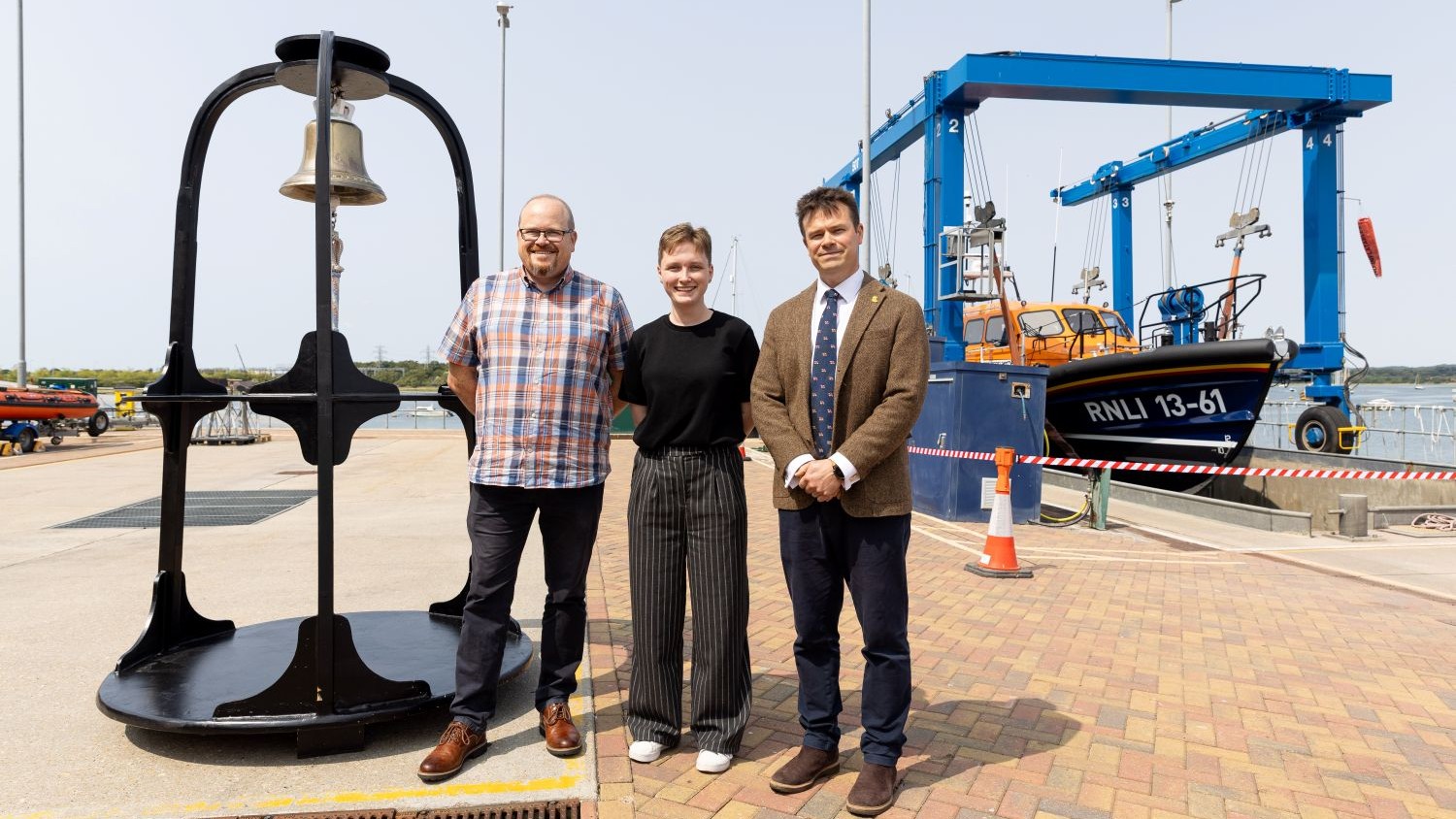 Three people standing smiling at the camera. Behind them is an orange all-weather lifeboat and to their left is a large brass bell hanging from a black frame