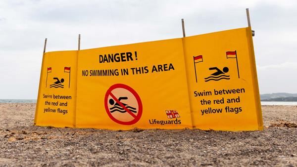 A warning message reading 'Danger! No swimming in this area' is written across the fabric of a yellow lifeguard windbreaker on a pebble beach