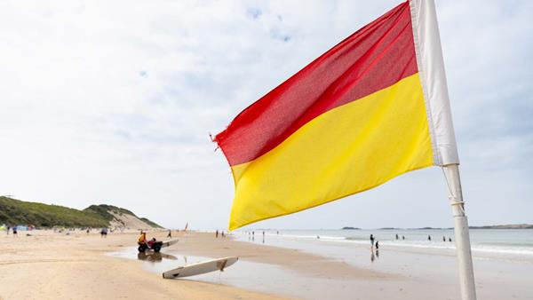 A red and yellow beach lifeguard flag flutters in the wind over a sandy beach on an overcast day