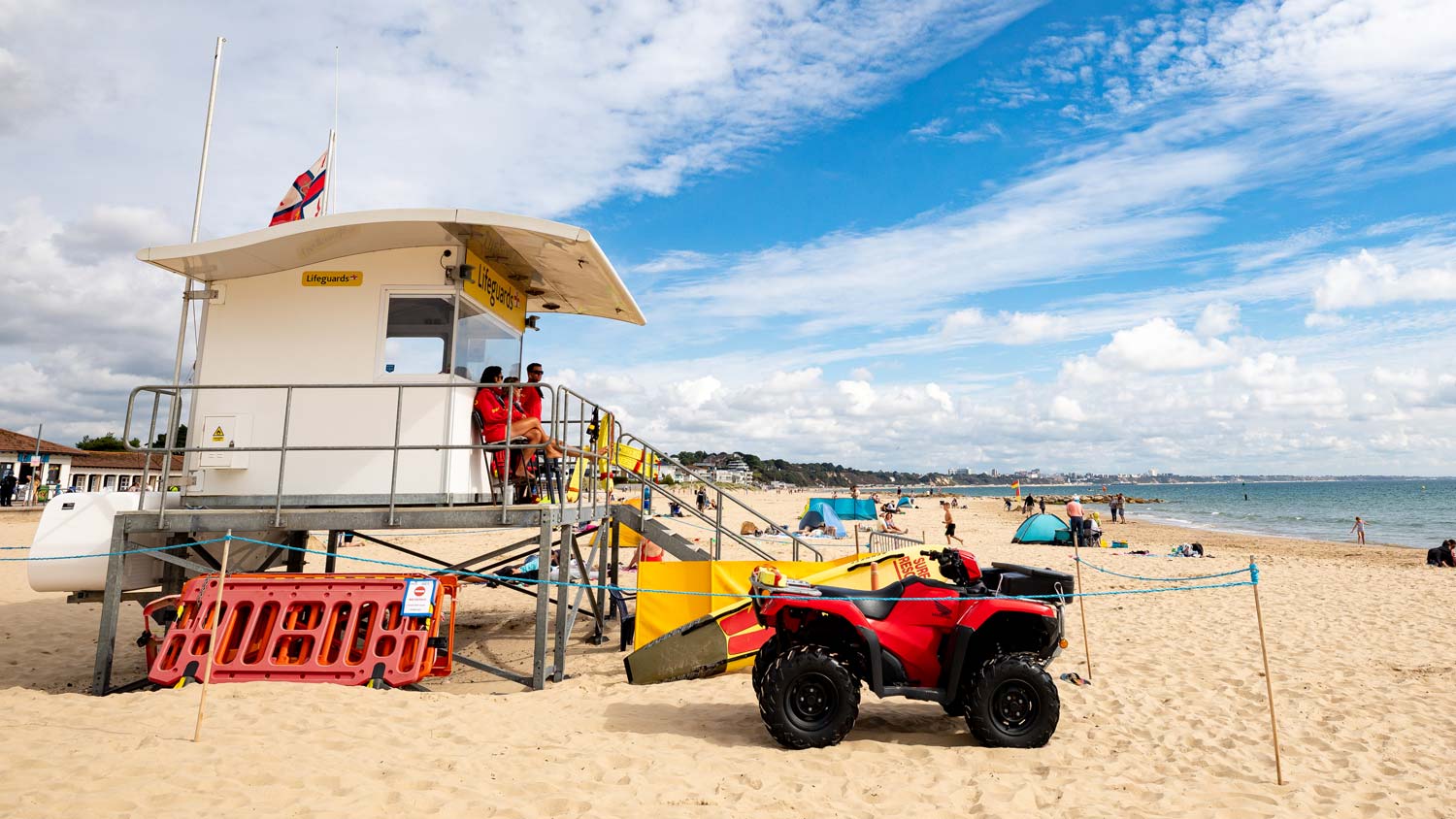 A trio of lifeguards in red jackets sit on the deck of their lifeguard hut keeping watch over beachgoers. A red beach ATV is parked on the sand beside it.