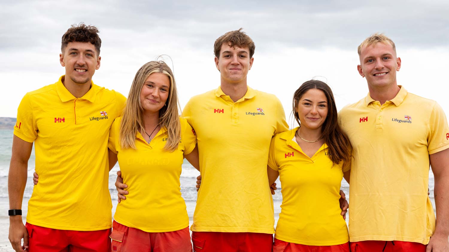 Group photo of Weymouth beach lifeguards featuring three male and two female lifeguards. They are wearing yellow lifeguard polo shirts.