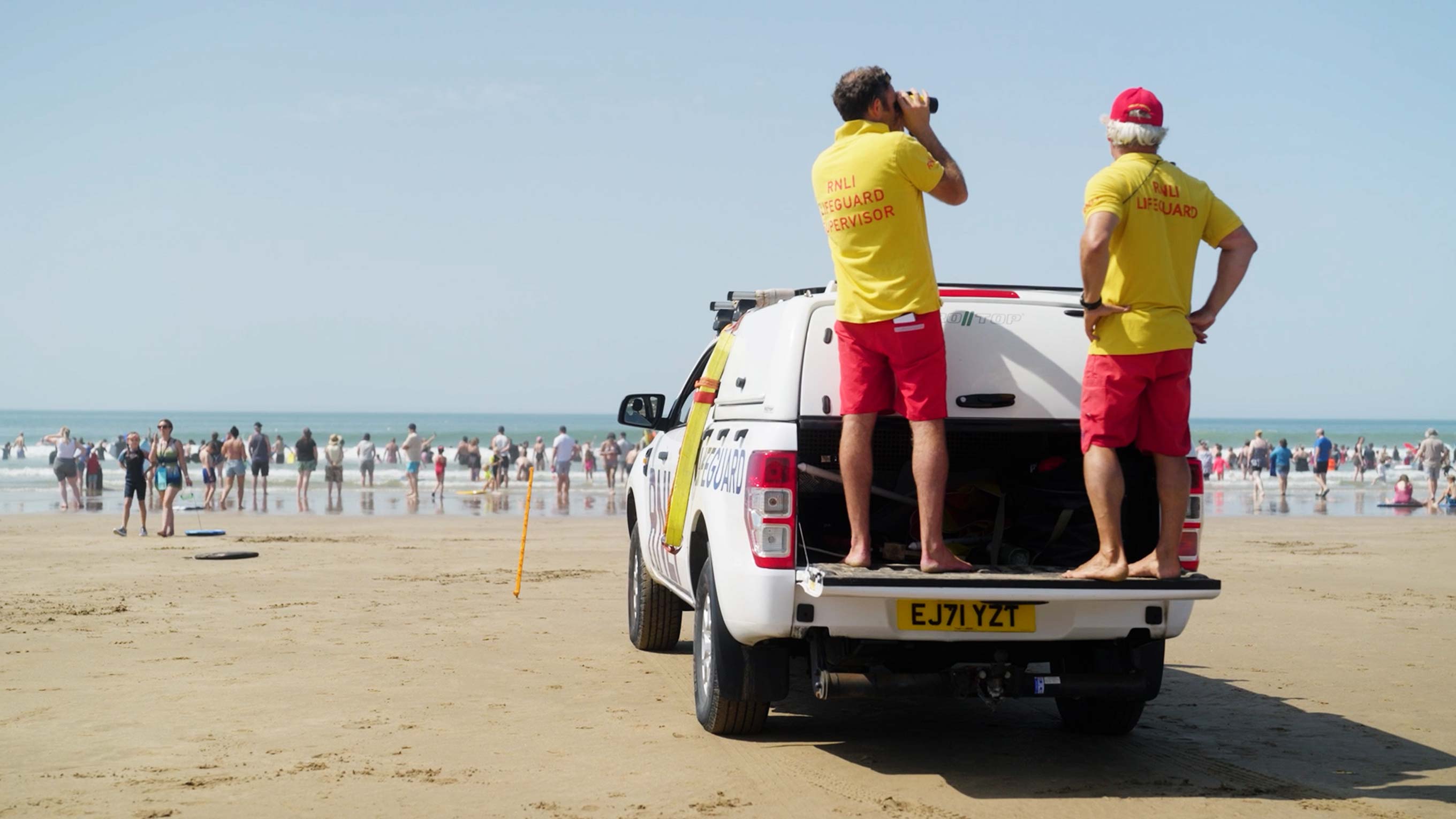 Two RNLI lifeguards wearing yellow shirts and red shorts stand on the back of a beach lifeguard SUV and survey the busy beach