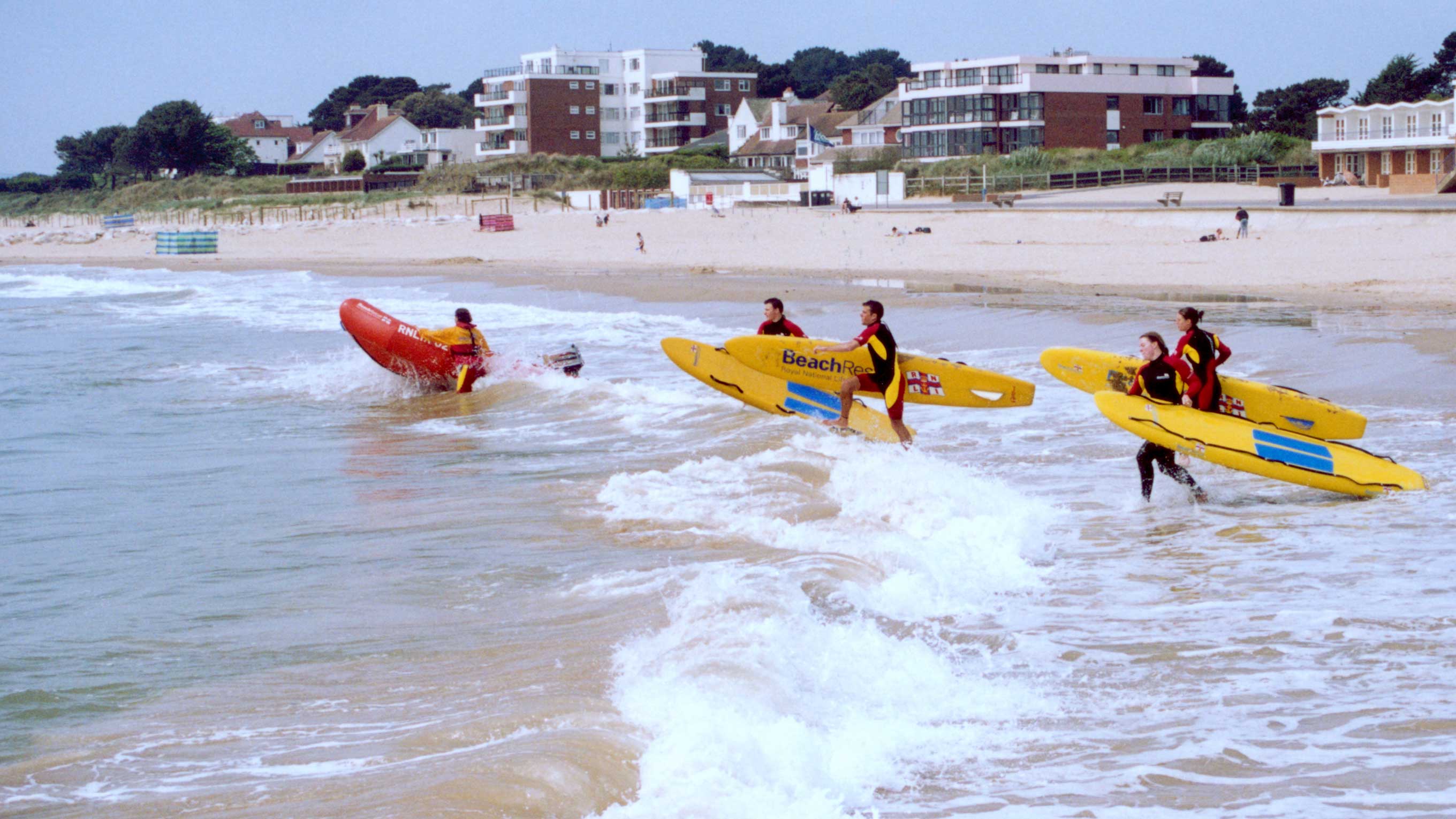 A team of RNLI lifeguards run into the surf carrying bright yellow rescue boards as part of a training exercise. One team uses as inshore rescue boat to power into the waves.