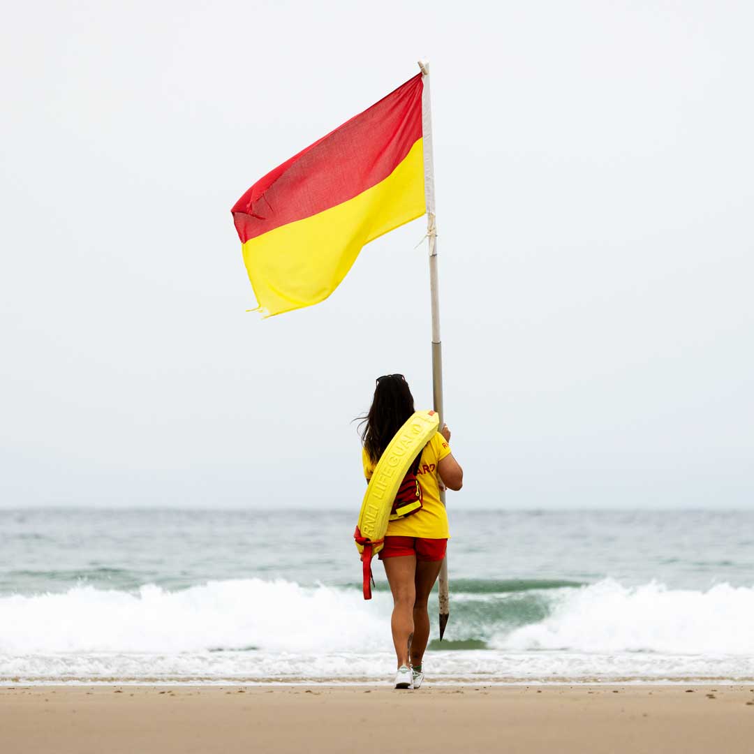 A female lifeguard carrying a red and yellow beach flag walks towards the surfline on a grey overcast day