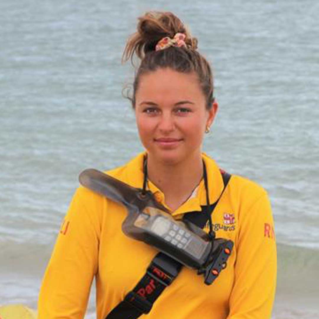 RNLI seasonal lifeguard Georgia Laing stands on the beach wearing yellow lifeguard polo and a radio strapped to her chest