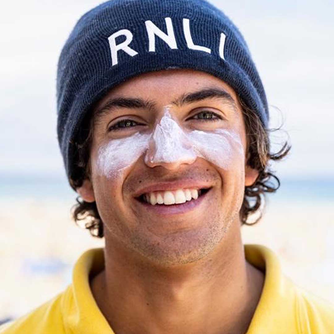 RNLI beach lifeguard Harry Barea with blue RNLI beanie and sunscreen on his smiling face