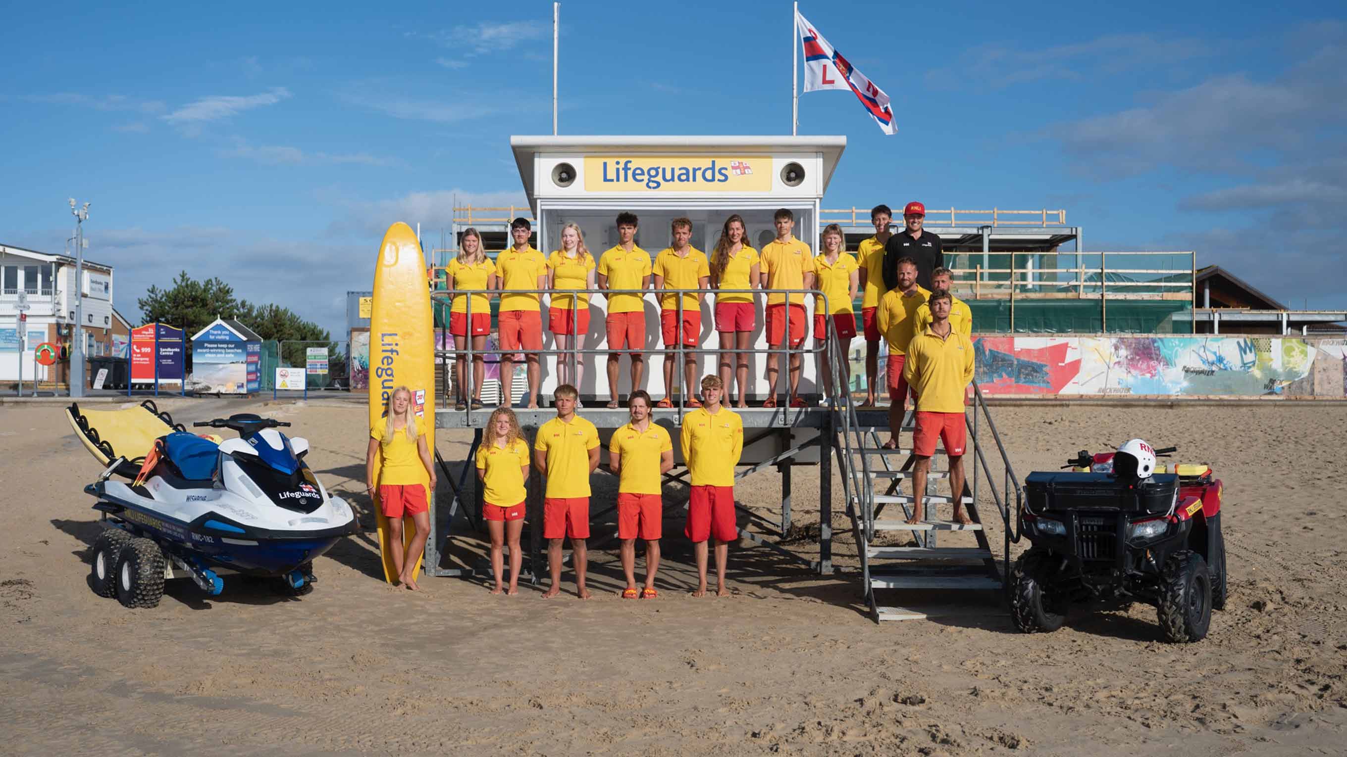 Group portrait of Poole RNLI beach lifeguards stood next to a lifeguard hut, rescue watercraft and ATV