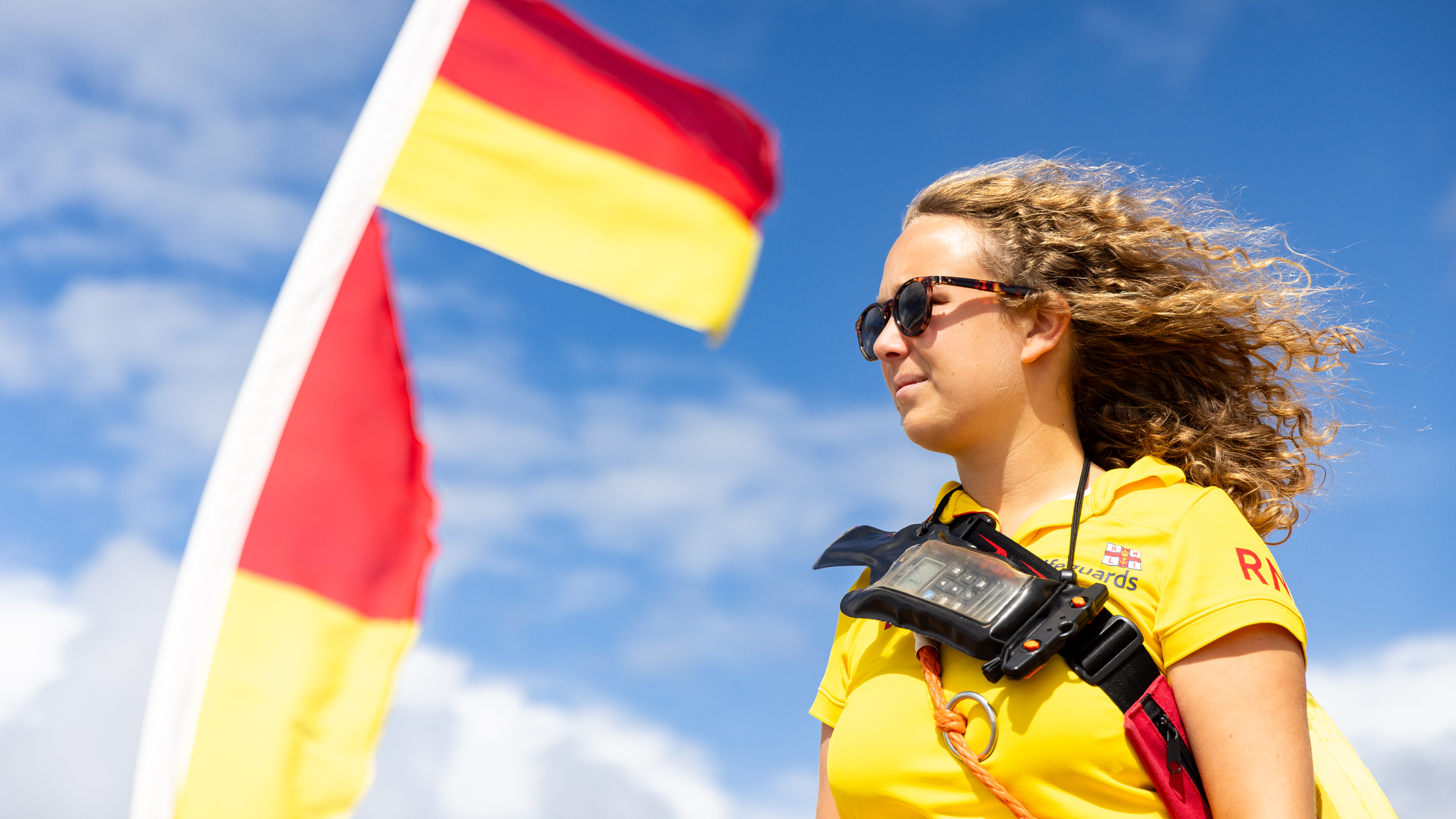 Lifeguarad Katie Stern stands on a beach next to a red and yellow beach flag watching the water. She is wearing a yellow polo and a radio strapped across her chest.