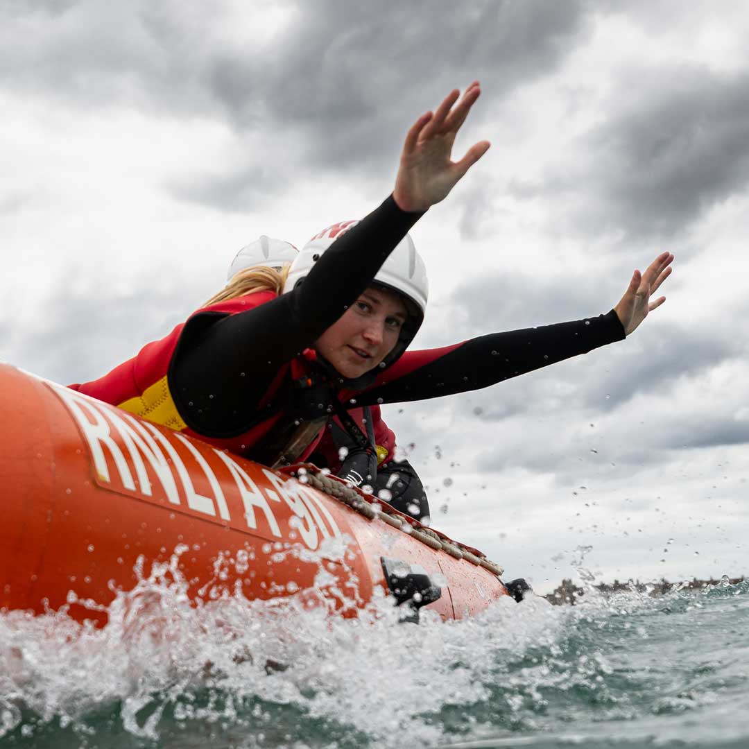 A female lifeguard in a wetsuit reaches out across the side of a RIB as it powers towards the camera