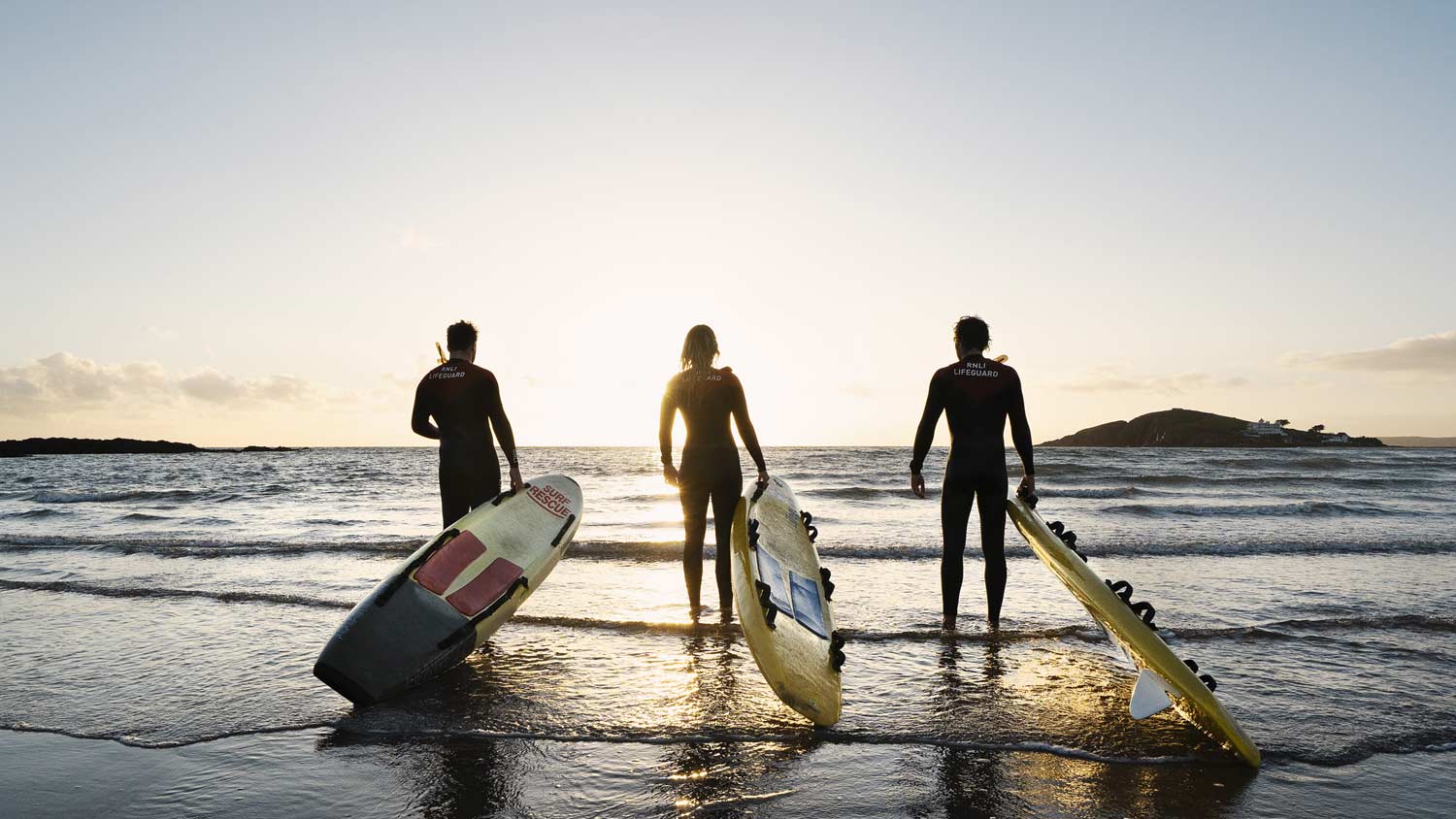 Three RNLI lifeguards holding rescue boards stand facing the sea as the sunsets over the horizon