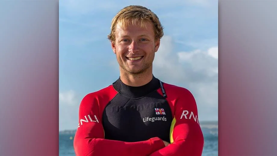 A male lifeguard smiles for the camera. He is wearing a red and black lifeguard wetsuit.