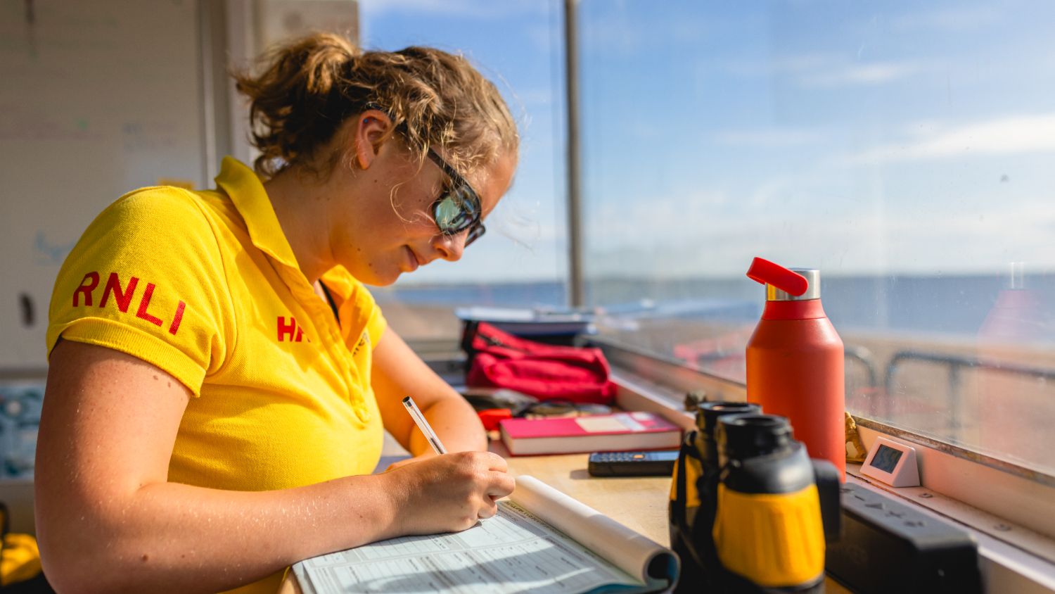 An RNLI lifeguard filling out paperwork inside the lifeguard hut at Broughty Ferry Beach, Scotland. She is wearing a yellow RNLI lifeguard polo, is wearing her hair in a ponytail and is also wearing sunglasses. There is other equipment on the table she is working at, such as a water bottle and a pair of binoculars.