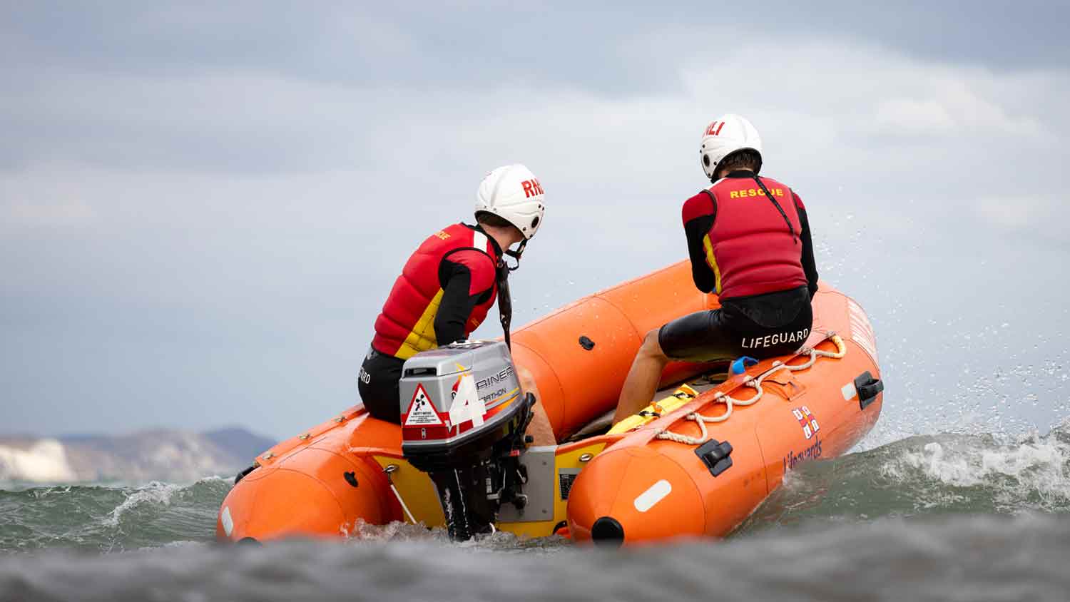 Two RNLI lifeguards wearing helmets, buoyancy aids and wetsuits sit onboard an inshore rescue boat as it powers over grey seas