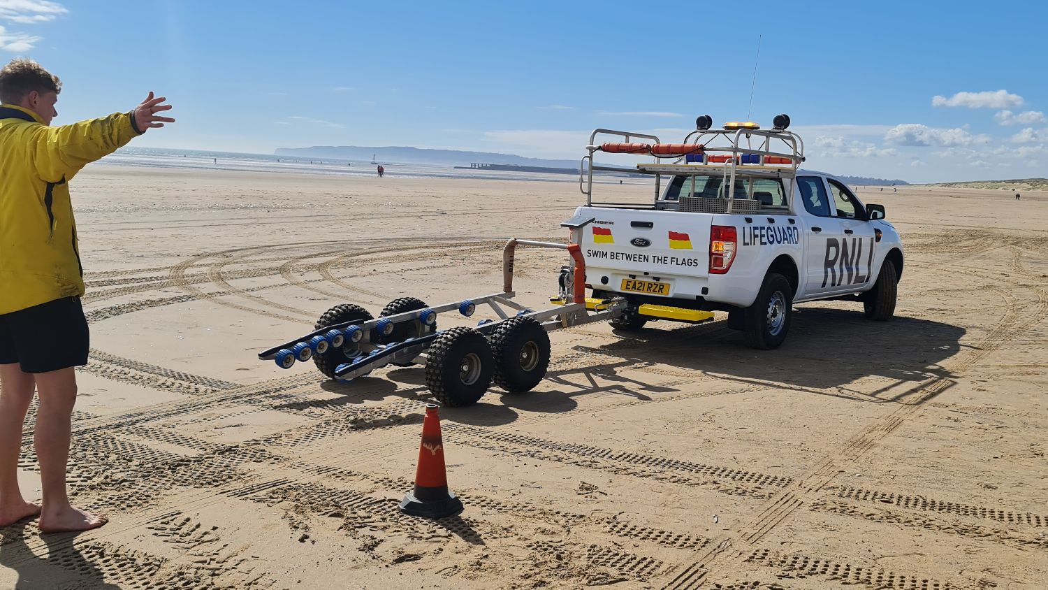 RNLI lifeguards taking part in four-wheel drive training using a patrol vehicle. They are on the sand at Camber Sands, East Sussex. One lifeguard is outside of the patrol vehicle with his right arm out to the side, signalling to the lifeguard driving the vehicle. The sky is blue with very few clouds.