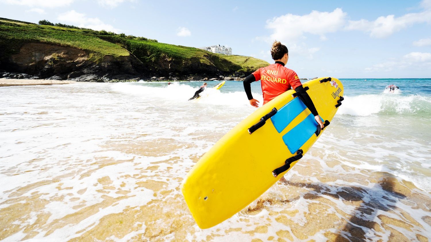 An RNLI lifeguard running into the sea holding a rescue board. There are other lifeguards in the sea also with rescue boards. They are taking part in lifeguard training at Poldhu Cove, Cornwall.
