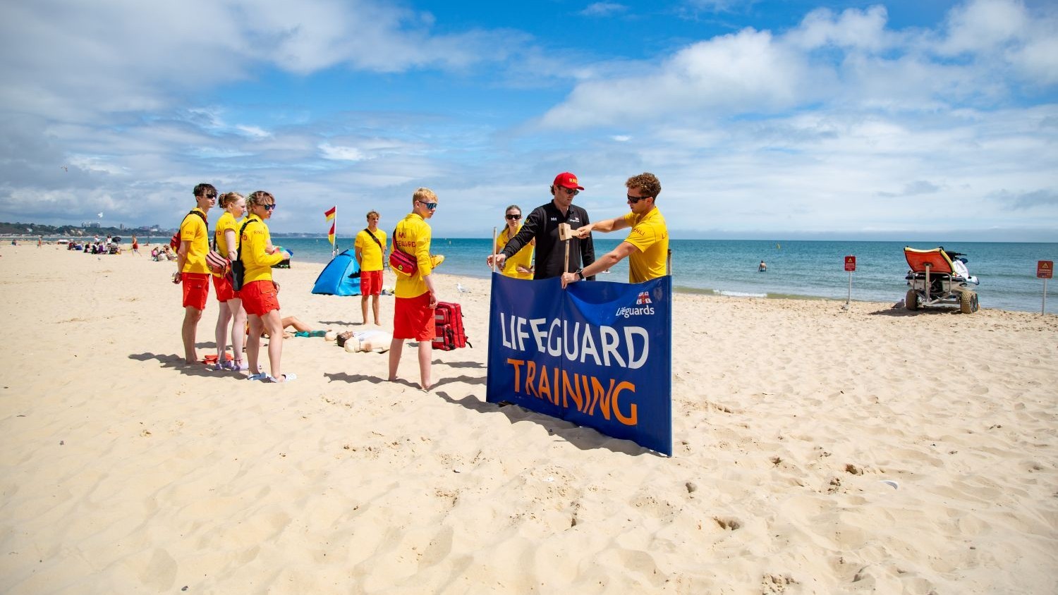 A group of RNLI lifeguards taking part in an induction on Sandbanks Beach in Poole, Dorset. One of the lifeguards is hammering a sign into the sand which says ‘Lifeguard training’ on it. The sky is blue with some clouds.