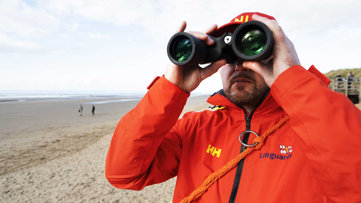 A lifeguard wearing a red lifeguards jacket stares out across the sea using binoculars