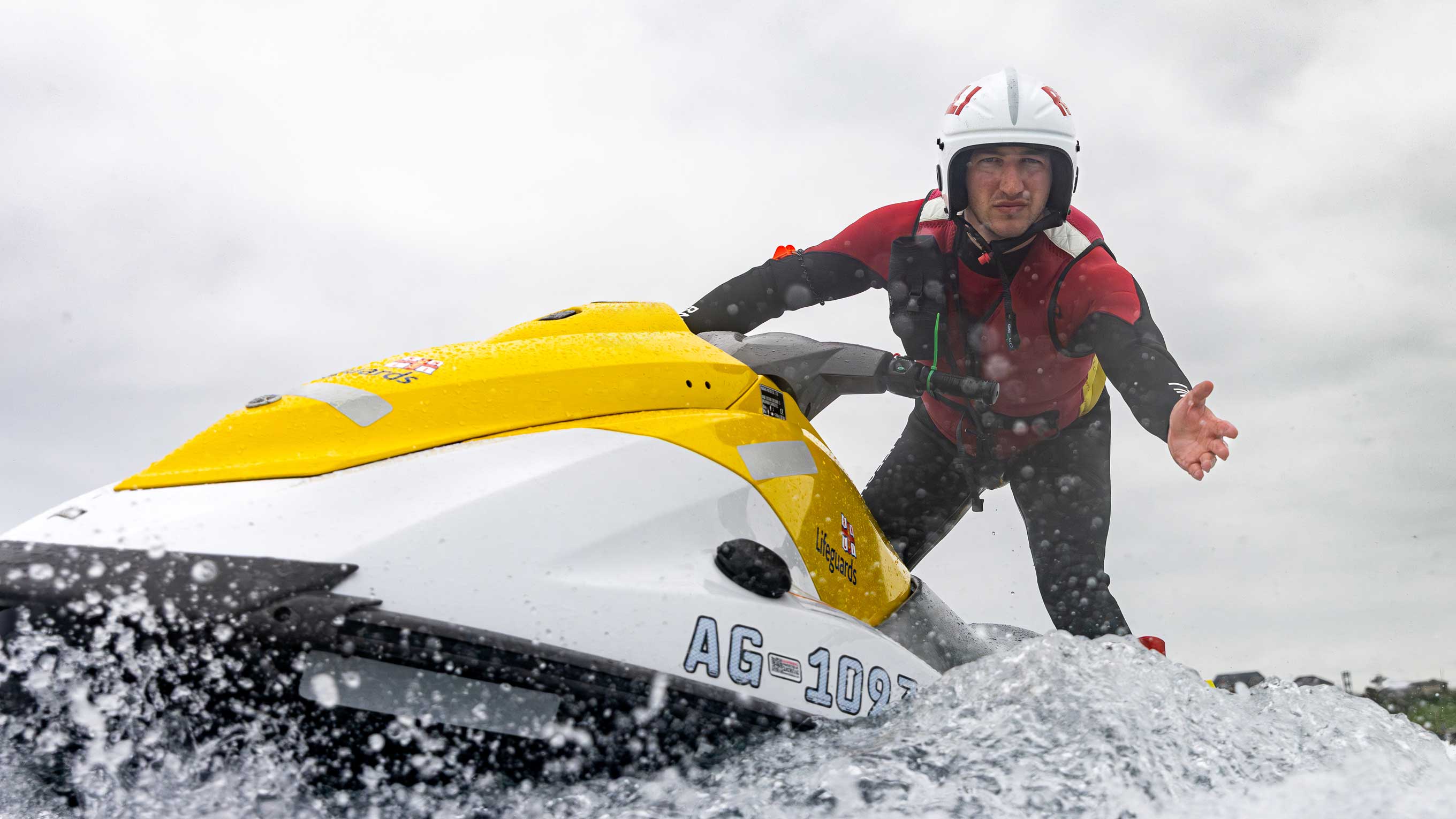 RNLI lifeguard supervisor Michael Jess reaches out towards the camera from onboard the back of a rescue water craft. He is wearing a red and black wetsuit, red rash vest and white helmet.