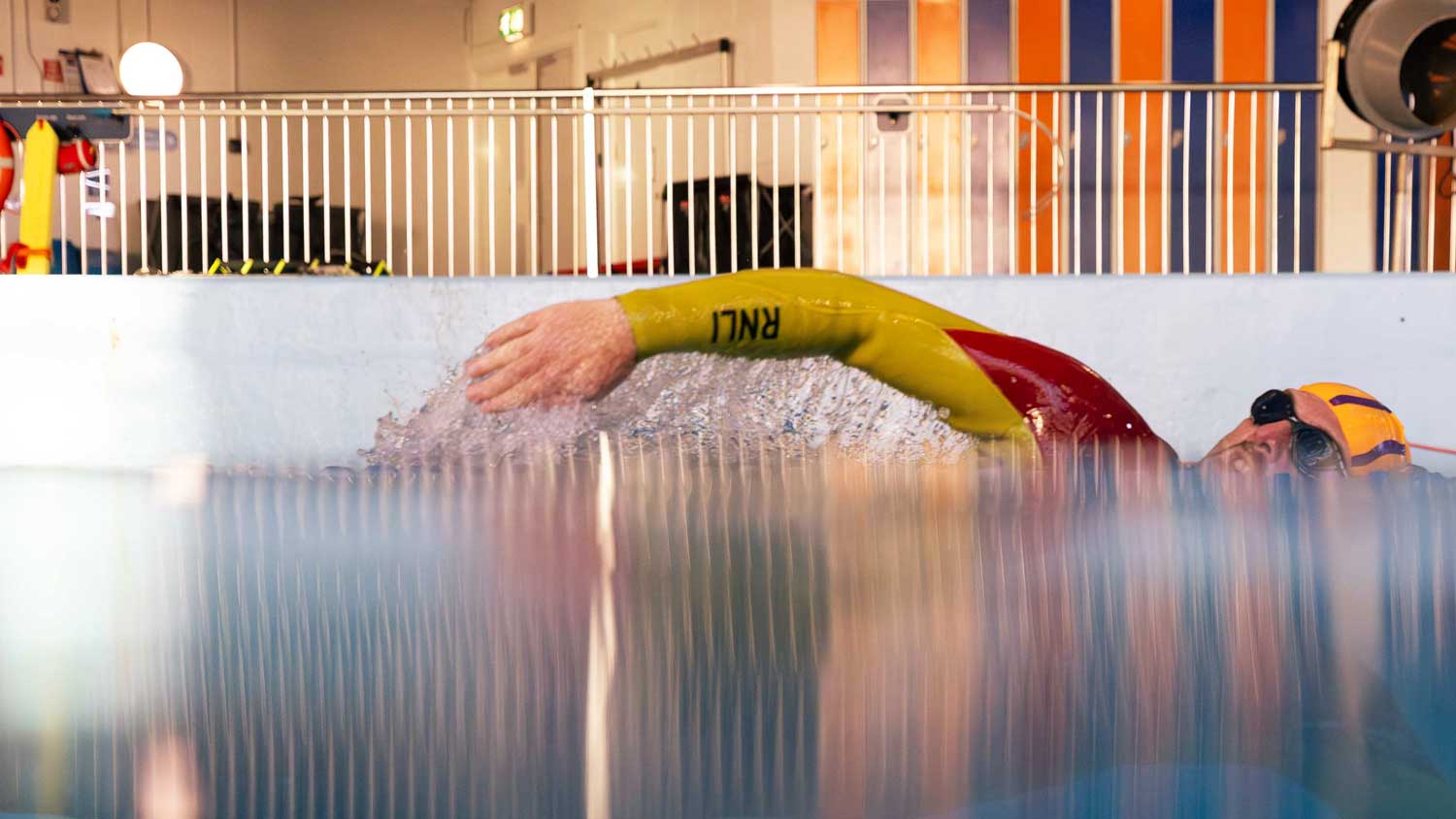 An RNLI lifeguard in red and yellow swimsuit, black googles and yellow swimsuit conducts a training swim through the RNLI sea survival pool