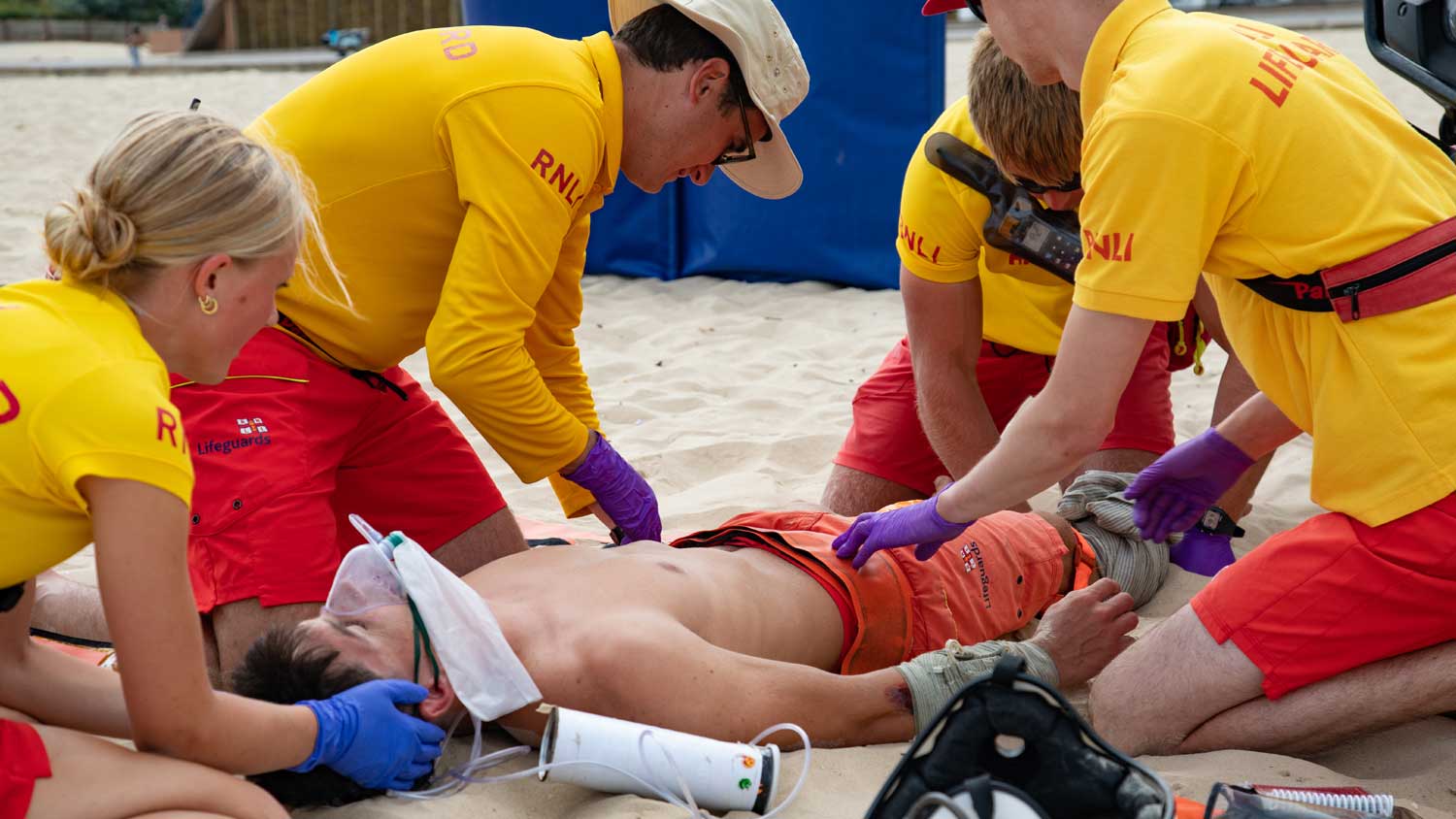 Four RNLI lifeguards carry out a casualty care training exercise on a  man. They are wearing yellow lifeguard t-shirts.