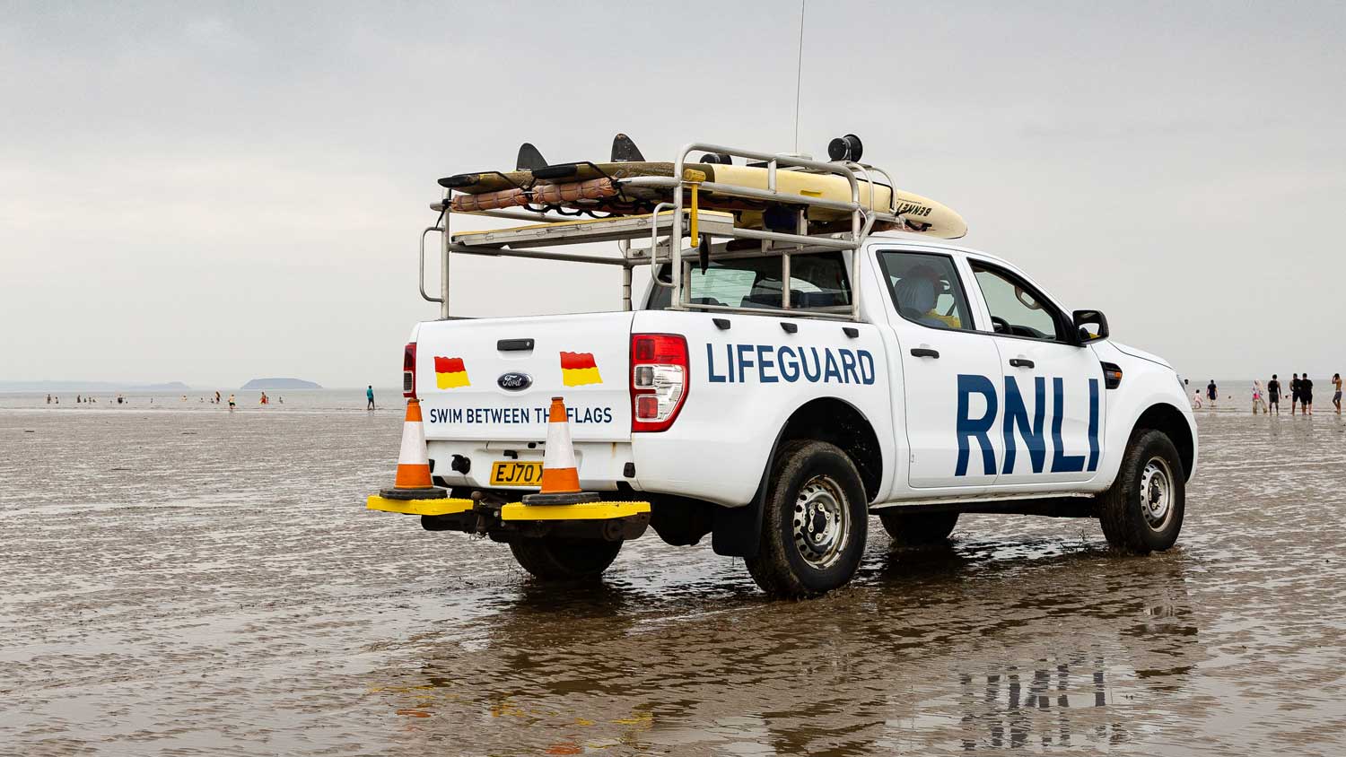 A white Ford lifeguard SUV sits parked on wet sands under grey skies