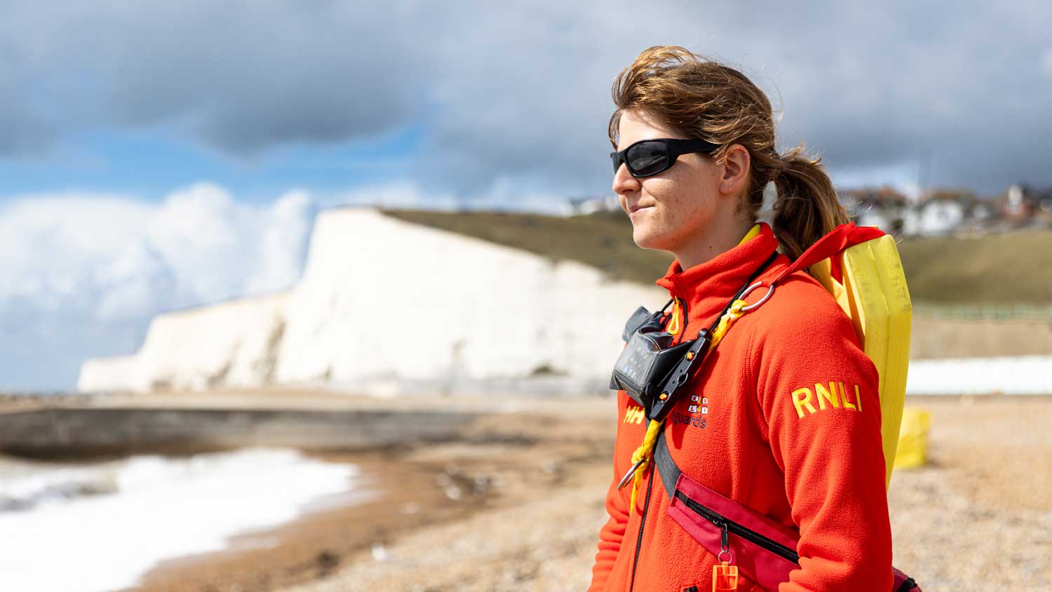 A female lifeguard wearing a red lifeguard jacket stares out to sea with white chalk cliffs behind her. She is wearing sunglasses, has a radio strapped across her chest, and a bright yellow rescue float strapped to her back.