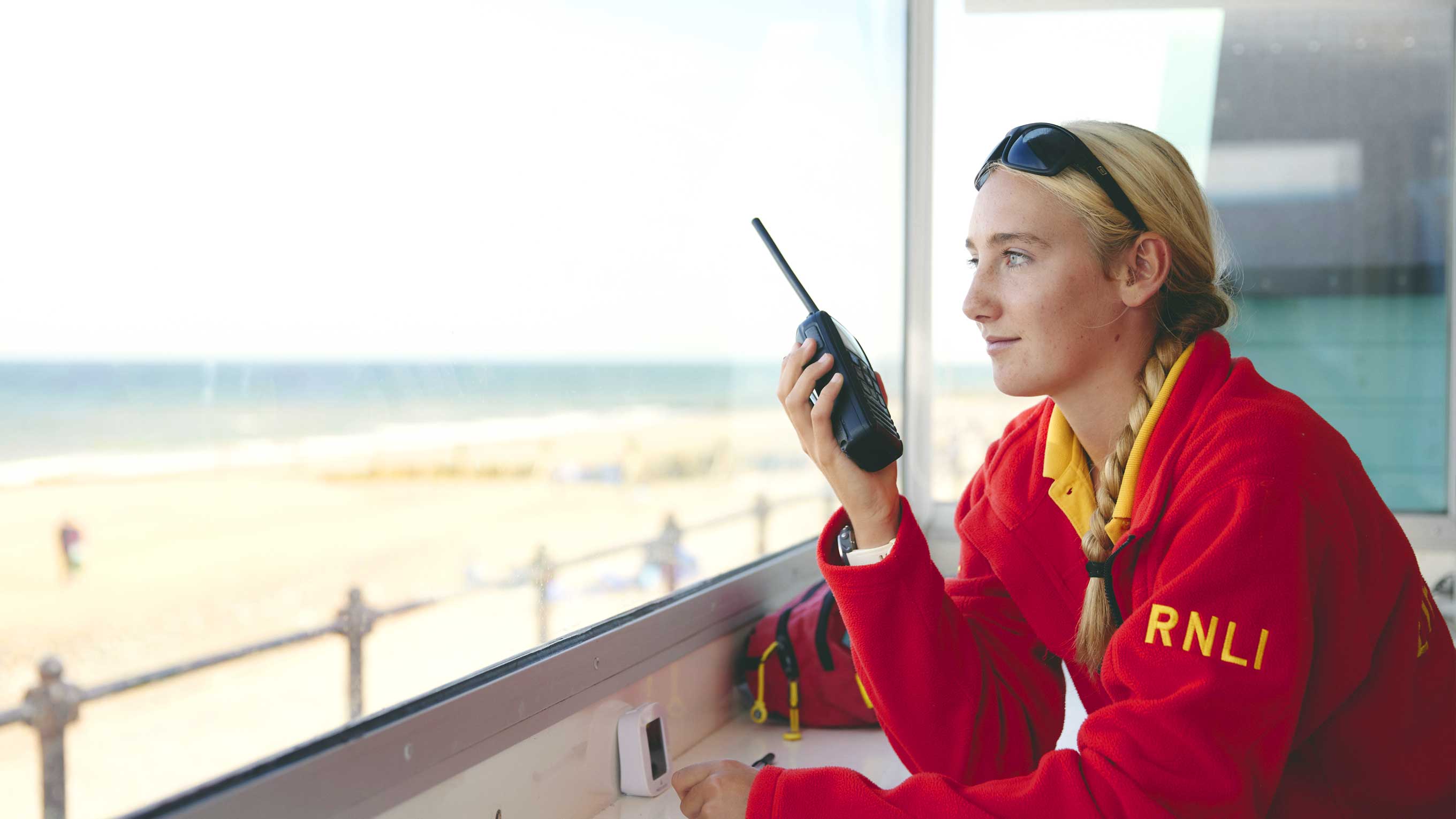 Lifeguard Grace Monsey sits inside a lifeguard hut watching over the beach. She is wearing a bright red lifeguard jumpet and holds a radio in one hand, ready to communicate.