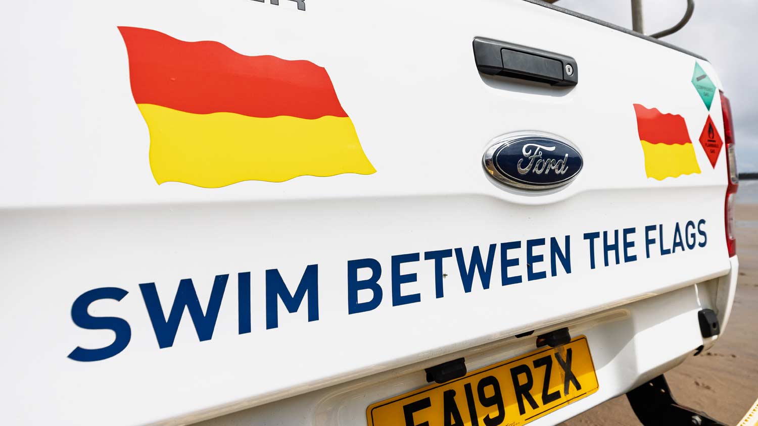 Close up of the Ford badge on the back of an RNLI lifeguard vehicle. Text either side of the badge reads 'Swim between the flags' and features graphics of red and yellow lifeguard flags