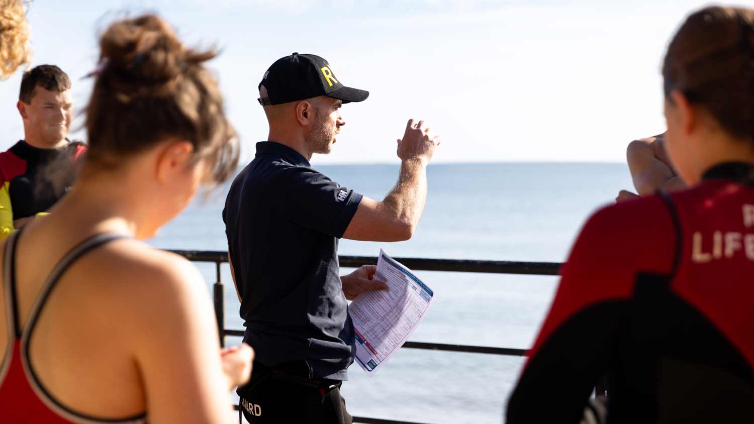 Lead Lifeguard Supervisor Andrew Jamieson briefing lifeguards at Scarborough North Bay beach. He is standing in front of railings pointing out to sea.