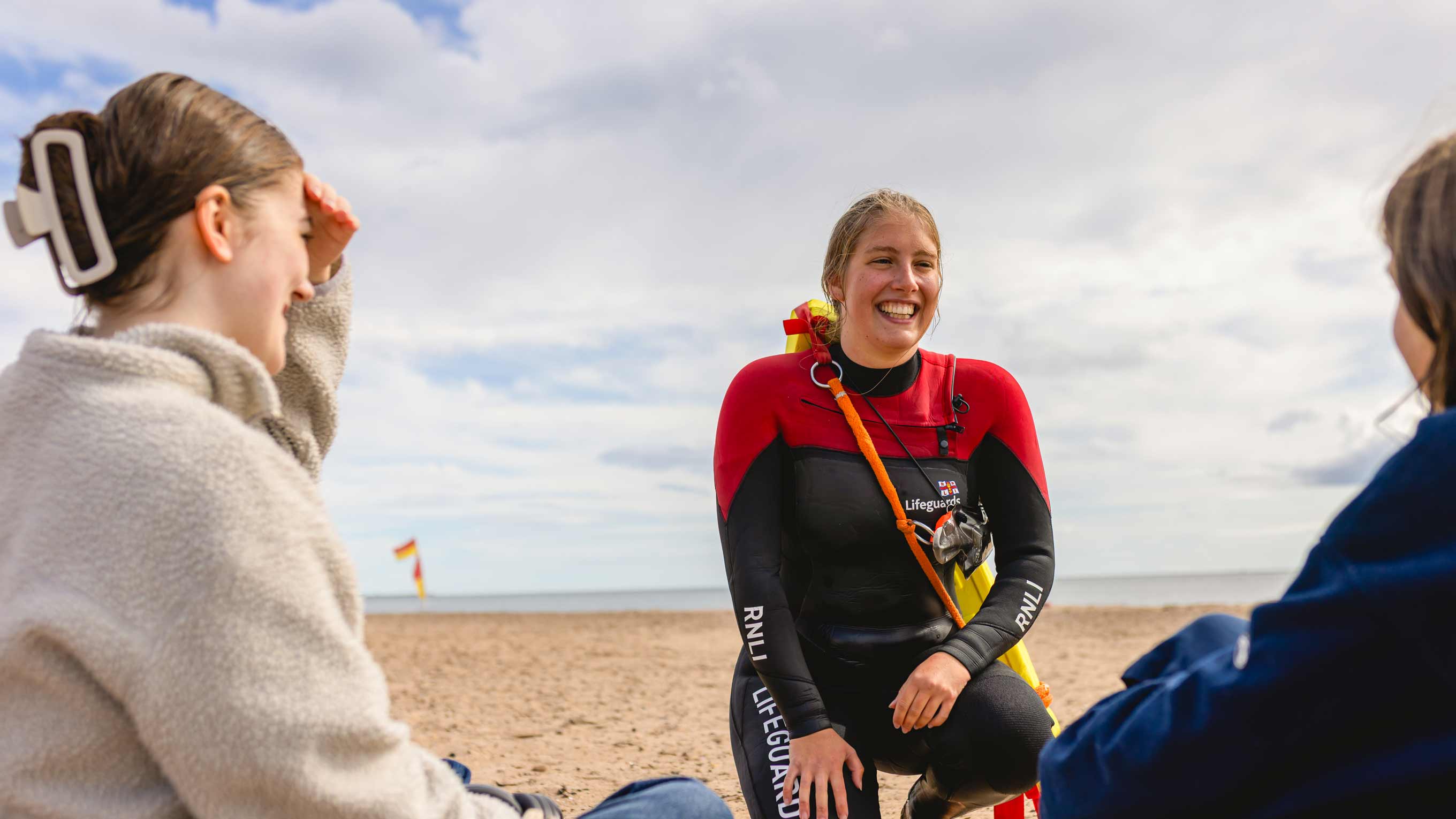 A female RNLI beach lifeguard in red and black RNLI wetsuit talks with members of the public