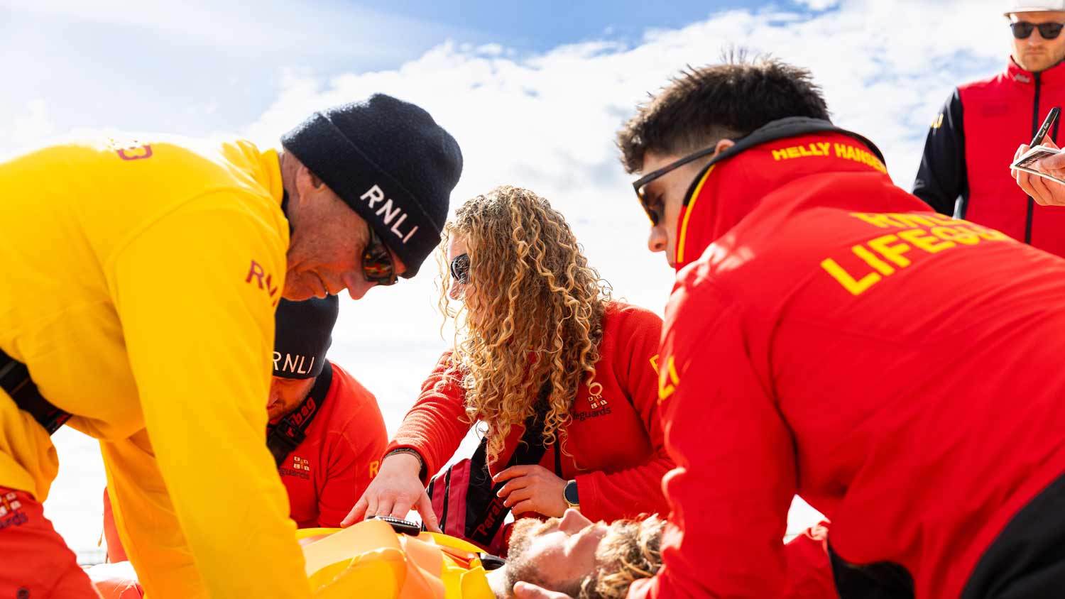 A group of RNLI lifeguards kneel around a person lying on the beach, supporting his head as they assess his condition, as they train in a casualty care scenario.
