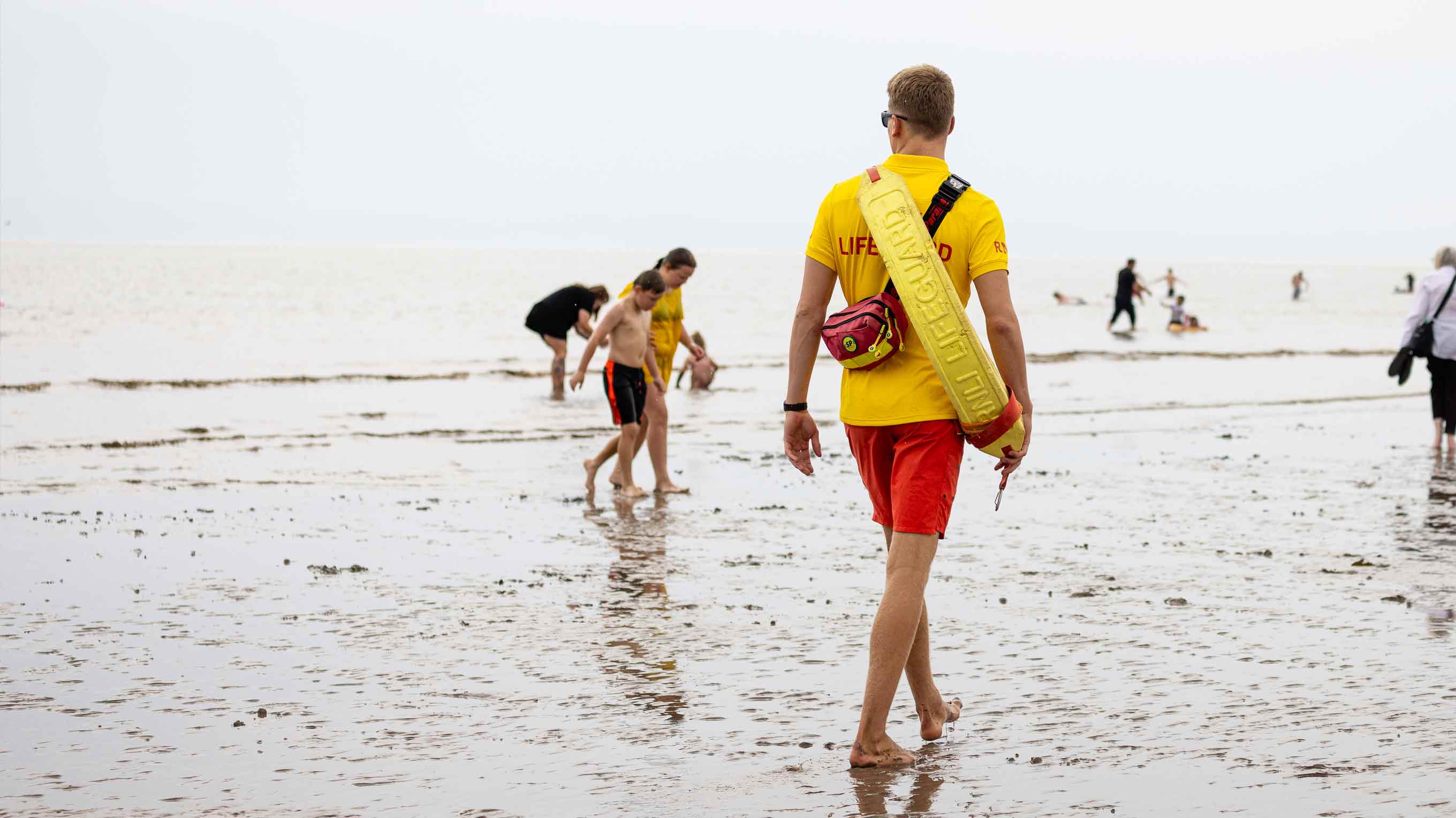 An RNLI beach lifeguard walks across wet sand while patrolling, wearing yellow shirt and red shorts, with a yellow lifeguard rescue float on his back. In front of him, beachgoers walk across the sand.