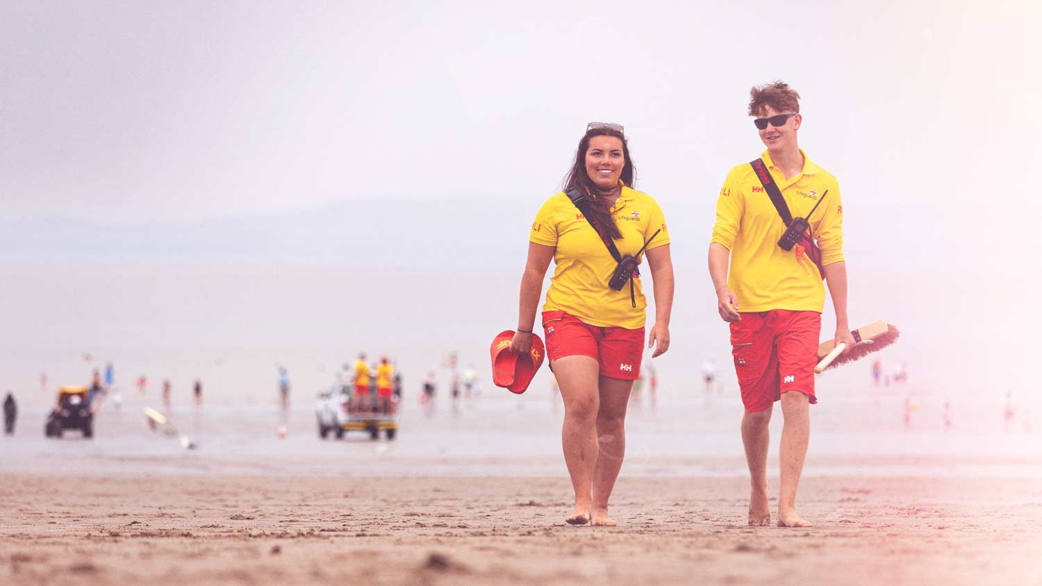 A pair of RNLI lifeguards in yellow polos and red shorts walk across the beach on patrol.