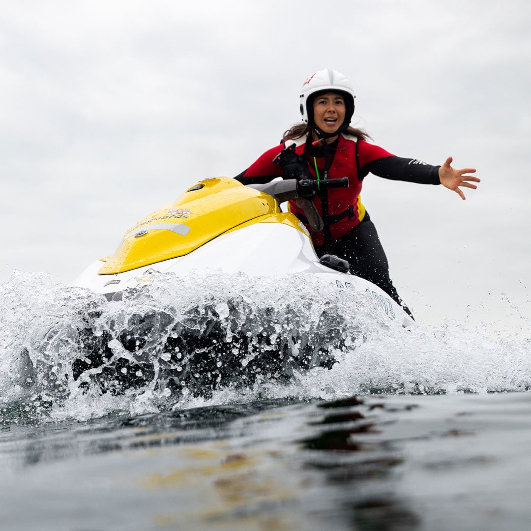 A female lifeguard wearing a helmet, buoyancy aid and wetsuit rides a rescue watercraft across grey seas. She reaches out towards a casualty off screen.