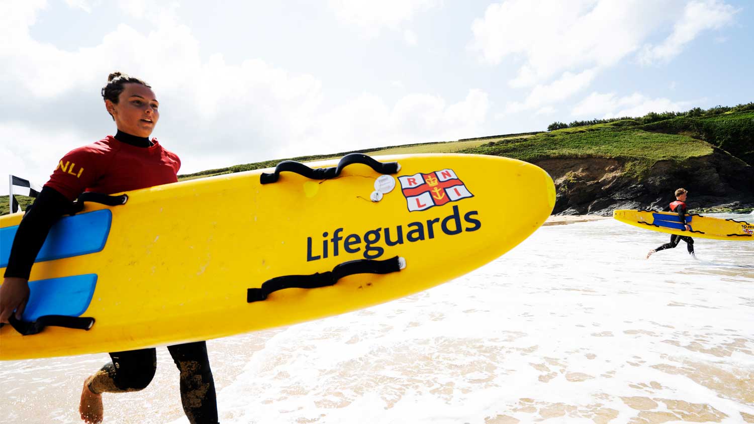 An RNLI lifeguard wearing a red and black wetsuit runs towards the surf line carrying a bright yellow lifeguards rescue board. Behind her, another lifeguard is also running into the surf.