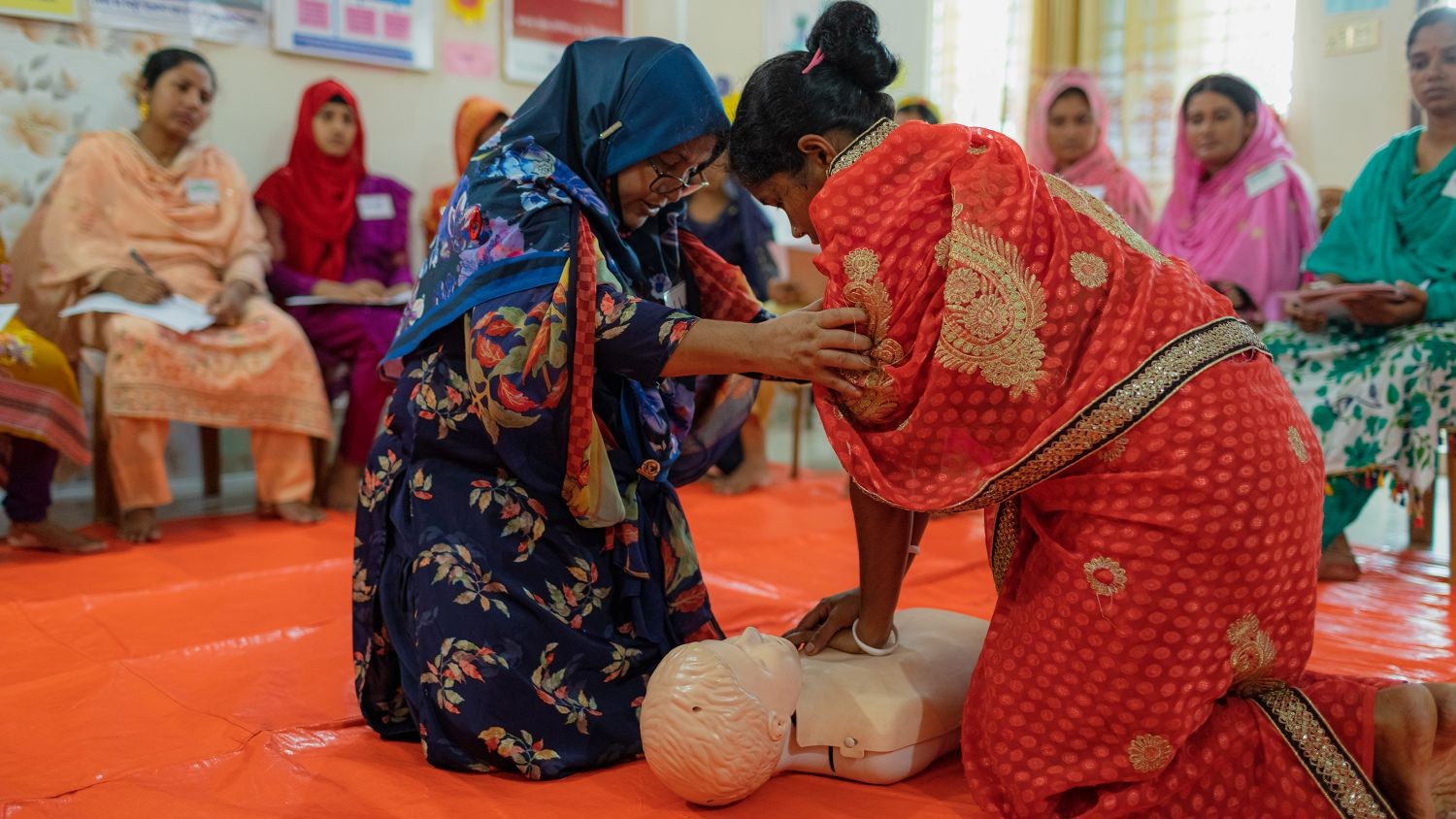 A woman is performing chest compressions on a first aid mannikin in a classroom setting. She is being guided by another woman. In the background, a row of women are seated, watching.