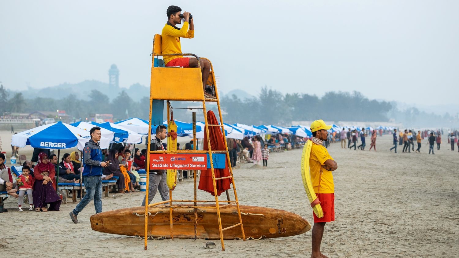 Two male lifeguards are watching over a busy beach in Bangladesh. One lifeguard is sat on a tall seat with binoculars, one is stood on the stand with his arms folded.