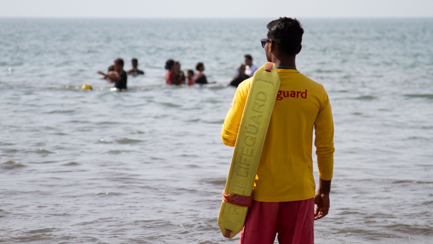 A lifeguard trained by the RNLI stands on a beach watching a group of swimmers in the water