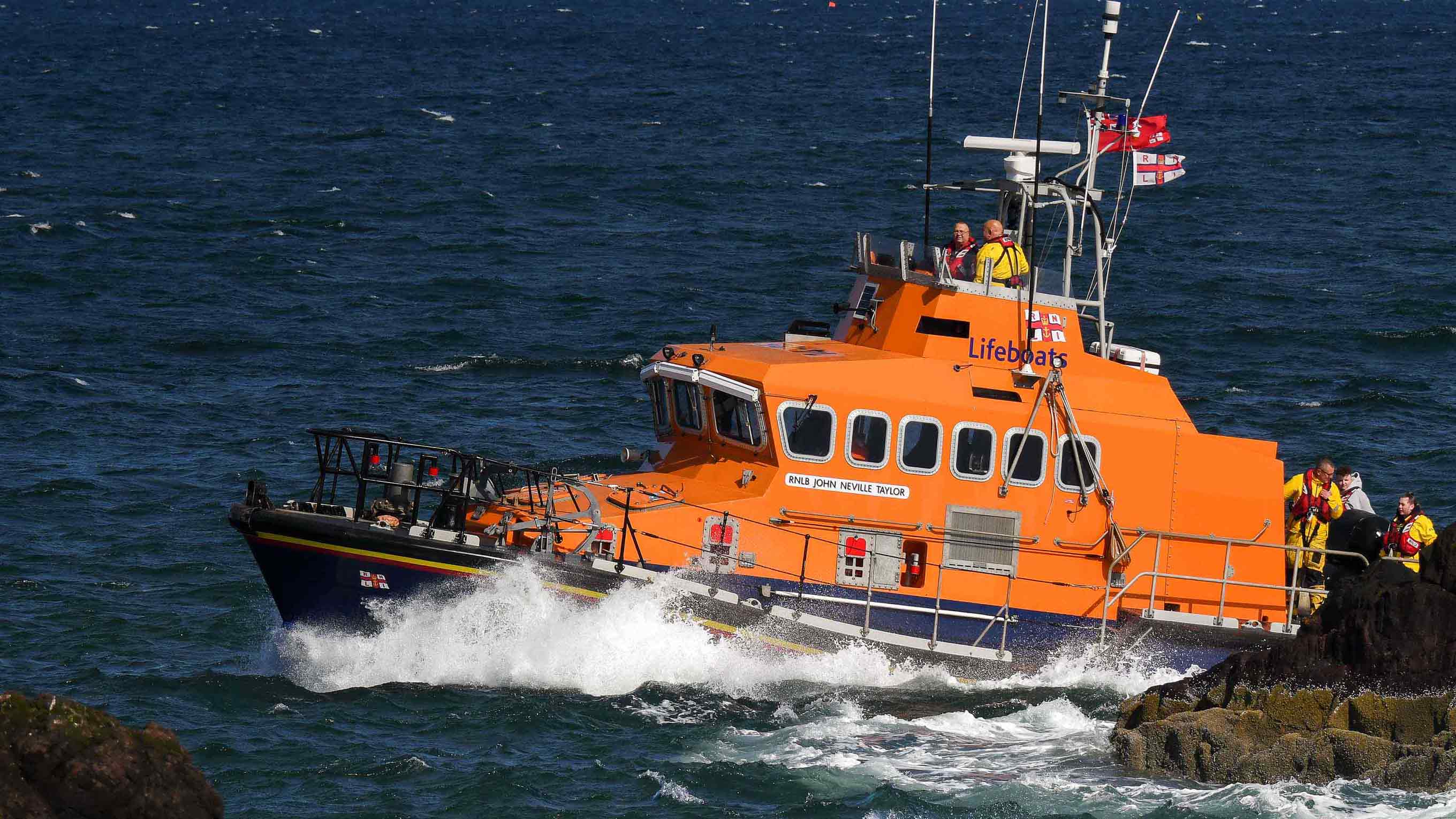Dunbar's Trent class all-weather lifeboat cruises along the sea past a rock formation. Two crew stand onboard the flying bridge with a further three on the rear deck. It's a bright sunny day.