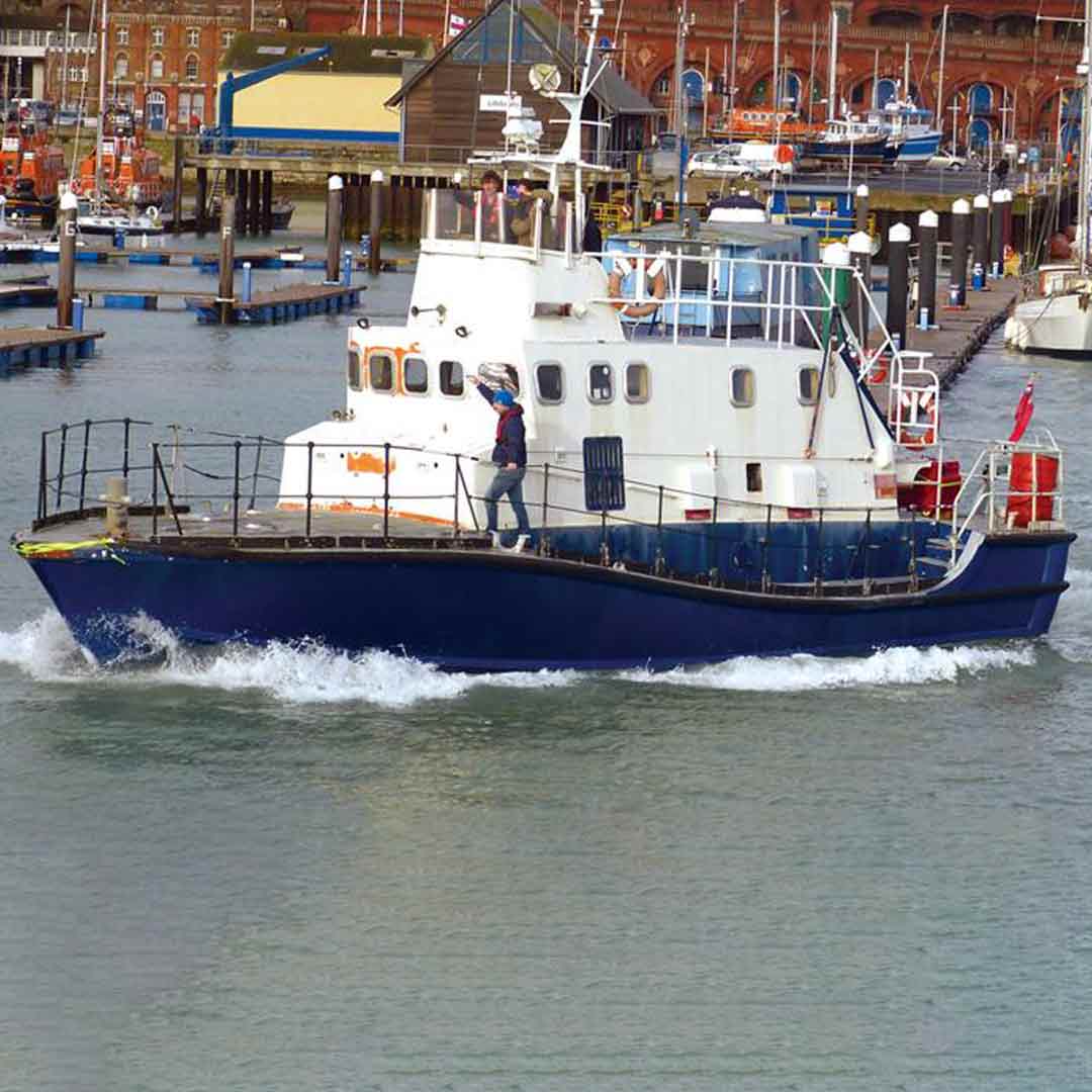 A white retired all-weather lifeboat cruises into a busy harbour. Onboard a crew member prepares the ex-lifeboat for docking.