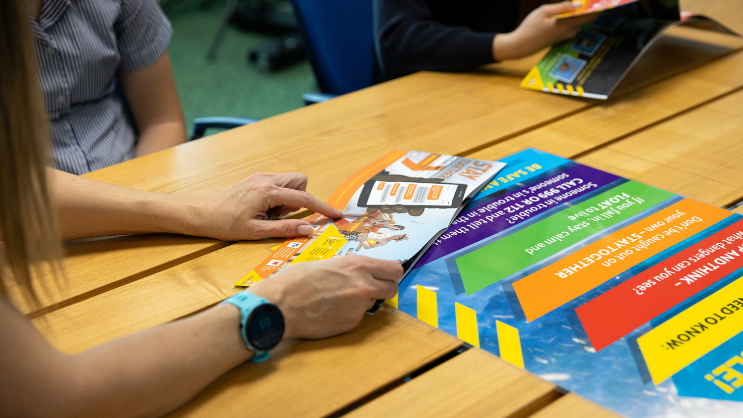 An RNLI water safety volunteer is looking through a brightly coloured booklet with a child, showing them tips about how to stay safe.  