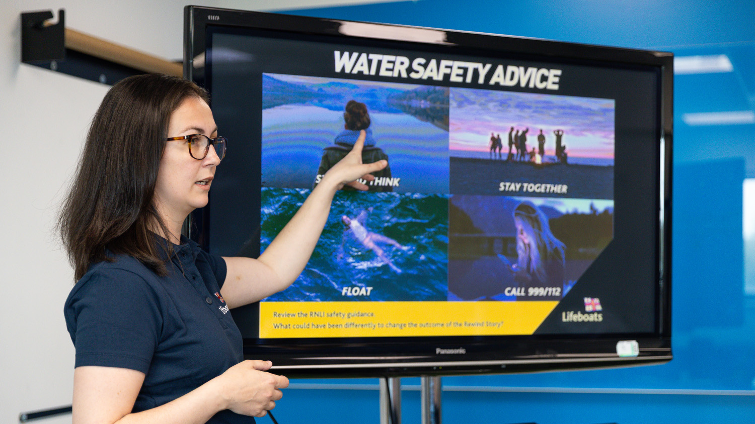 An RNLI water safety presenter is pointing to a television screen at the front of a classroom, showing the pupils vital water safety advice. 
