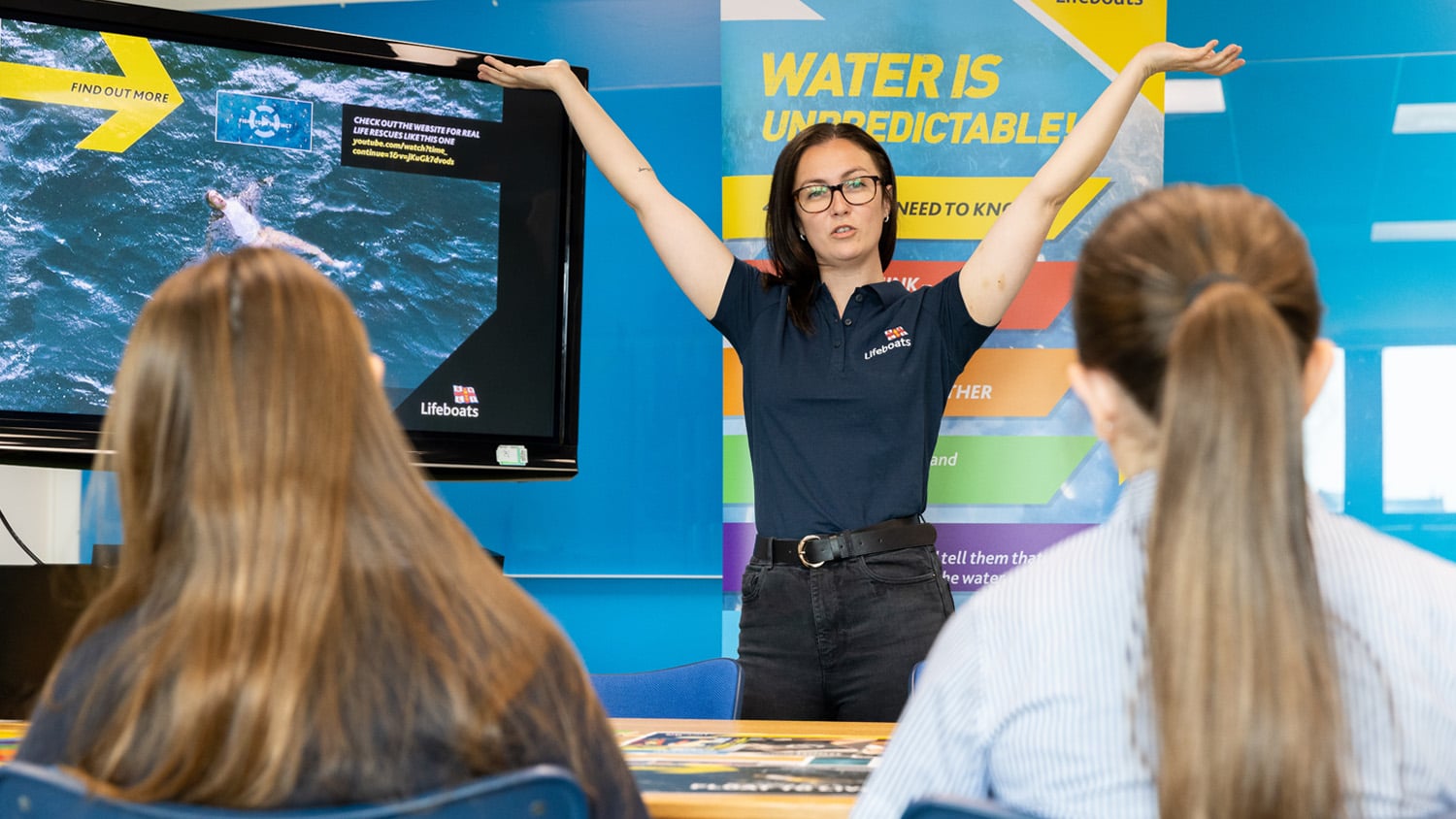 A water safety volunteer is standing at the front of the classroom, showing the students a Float to Live video. She has her arms raised in the air, showing them how to float. 