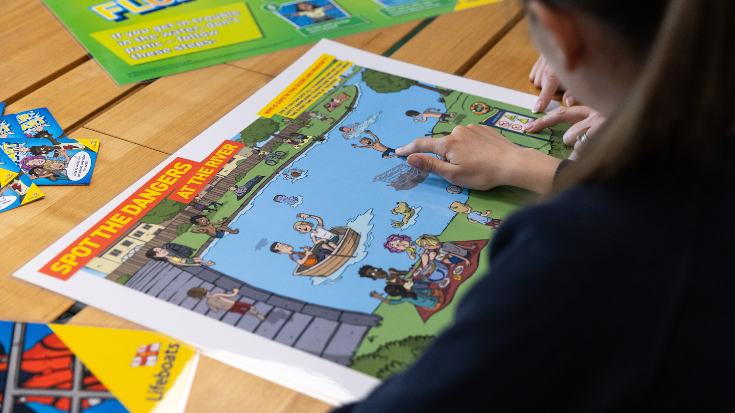 Schoolchildren are looking at an illustrated 'spot the dangers at the river' activity, and pointing to dangers in the water. 