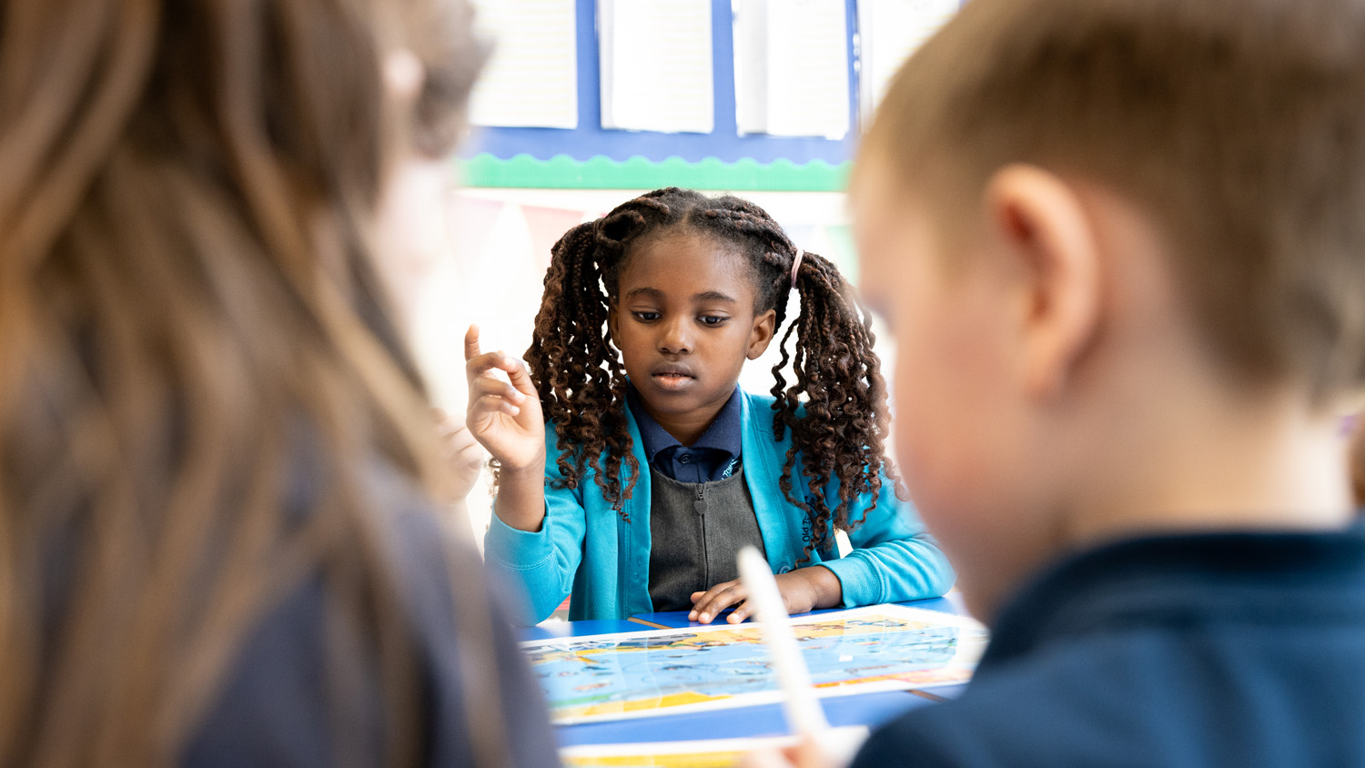 Schoolchildren are in a classroom, working on RNLI water safety activity sheets. One child has her finger pointing upwards, asking a question. 