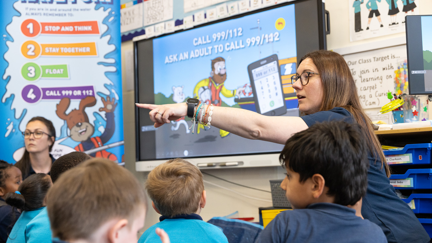 Two RNLI presenters are giving a water safety talk to children at an infant school. There's a roller banner and a digital screen behind them showing safety tips like floating and calling 999 or 112 for help.