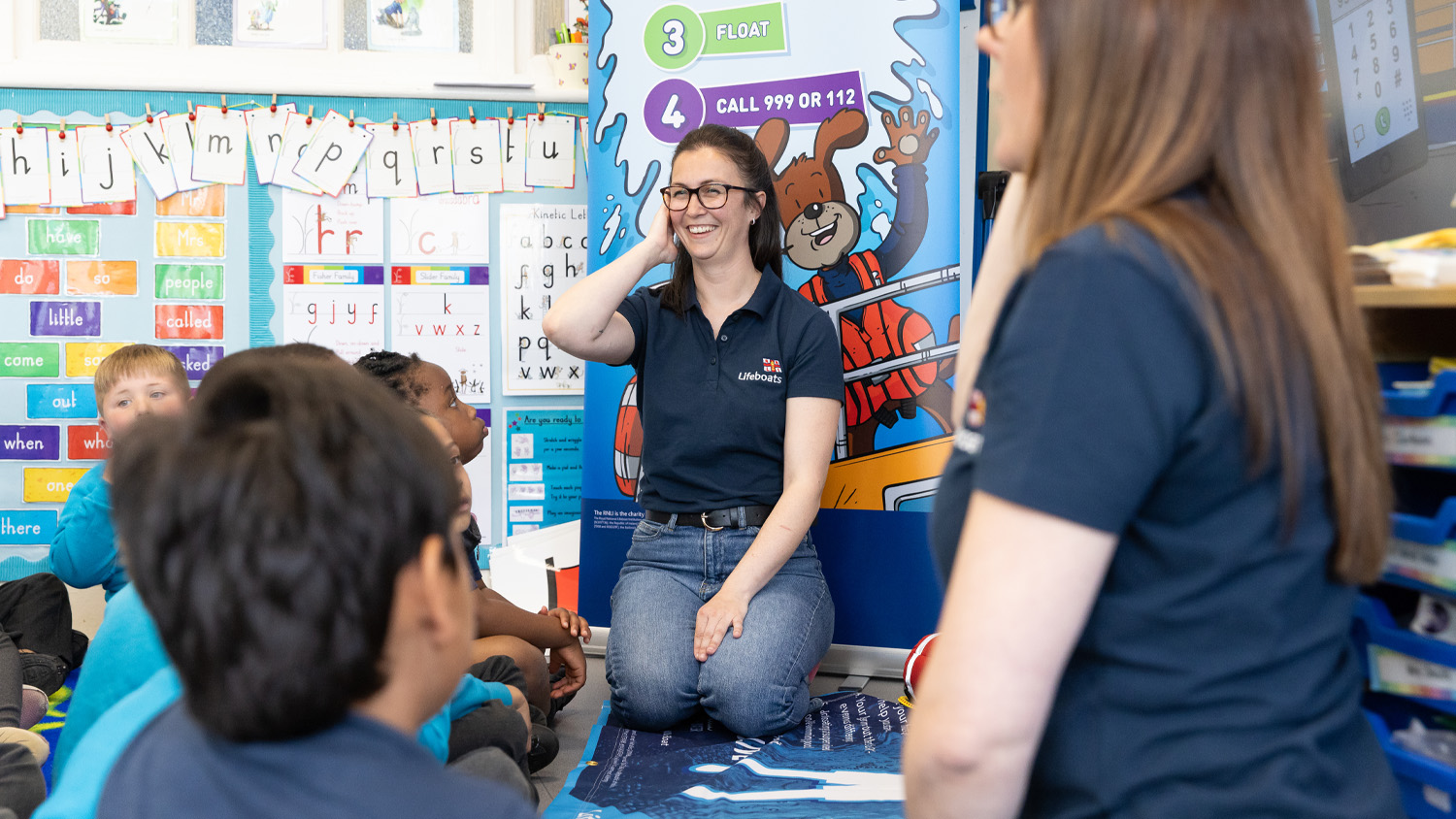 Two RNLI presenters are giving a water safety talk to children at an infant school. There's a roller banner behind them showing safety tips like floating and calling 999 or 112 for help.