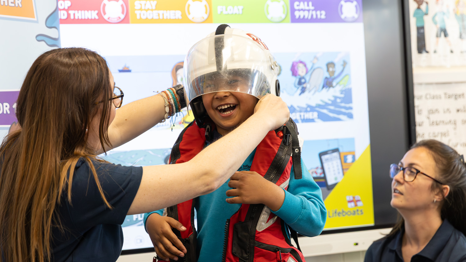 A young child is laughing while trying on crew kit, with an RNLI water safety presenter helping them put on a white helmet. 