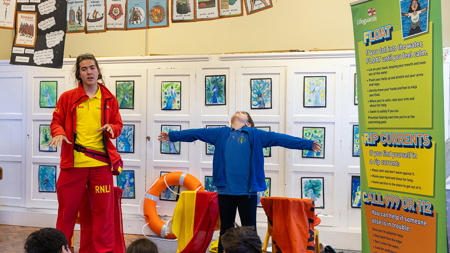 An RNLI lifeguard wearing a red fleece and trousers stands in front of a group of school children at a meet the lifeguard event. Beside him, a schoolchild wearing a blue fleece and black trousers extends their arms out as they demonstrate the float position.