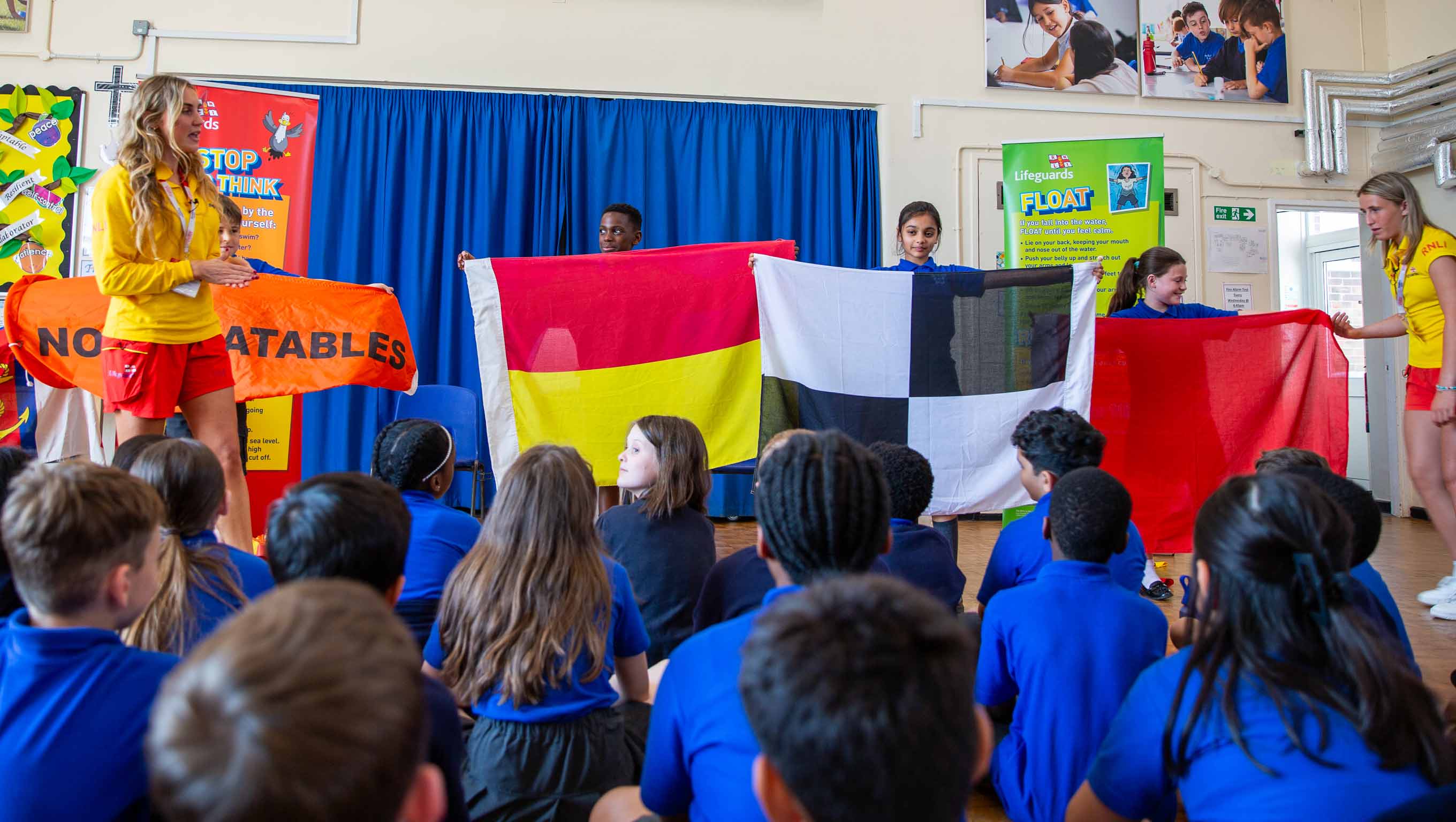 Two female RNLI lifeguards wearing yellow polo shirts and red shorts present at a school assembly. A group of school children stand between them holding the different types of beach flags as children sit on the floor and watch